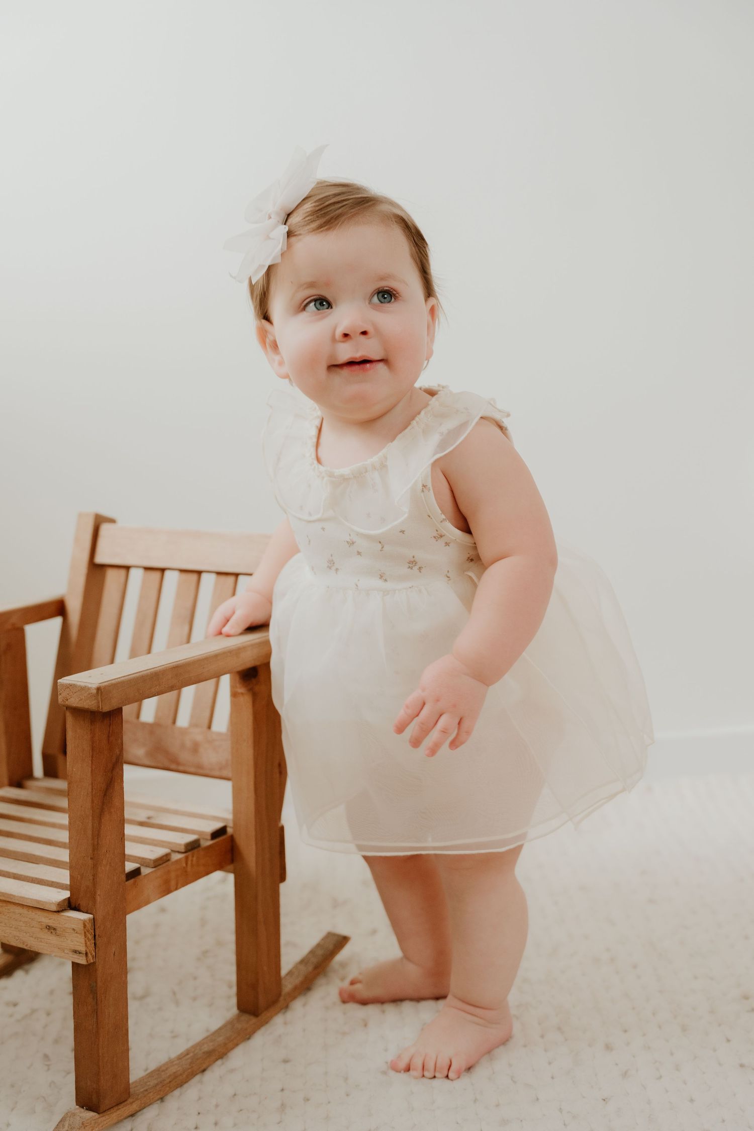 A baby in a white dress poses near wooden furniture against a light background in a series of connected photos.