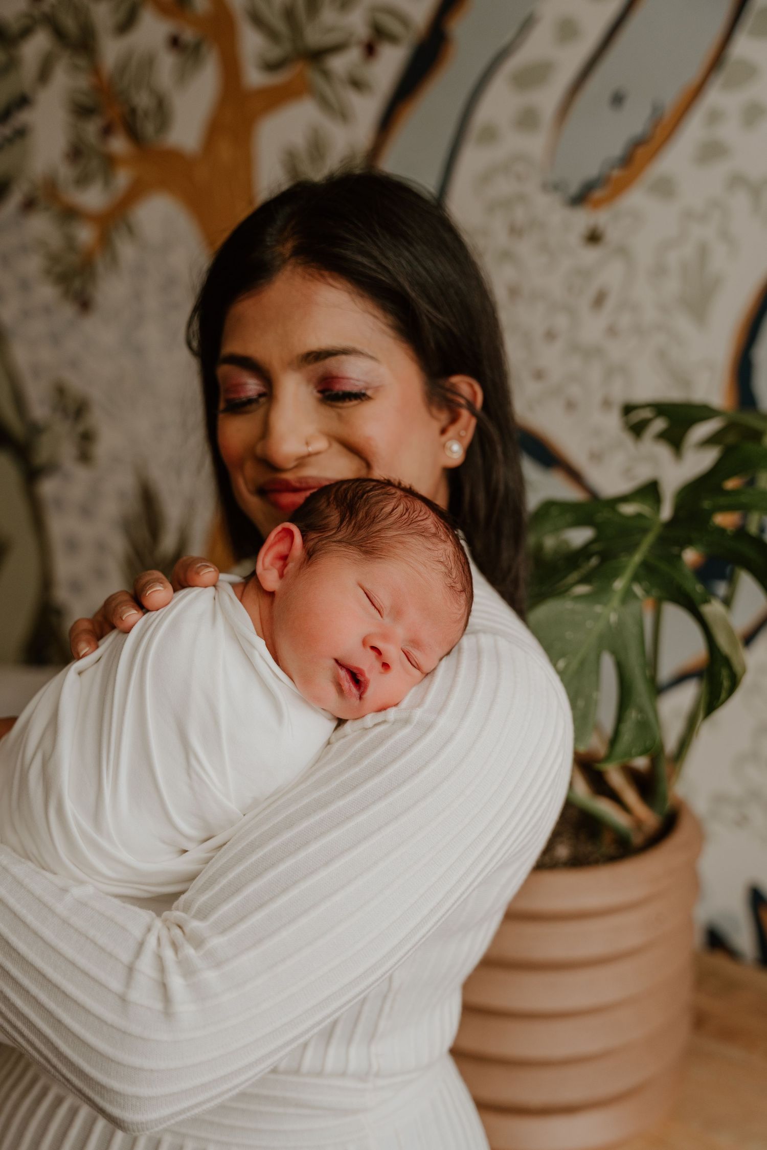 A tender moment captured as a sleeping newborn rests peacefully against someone wearing a white sweater beside a potted plant.