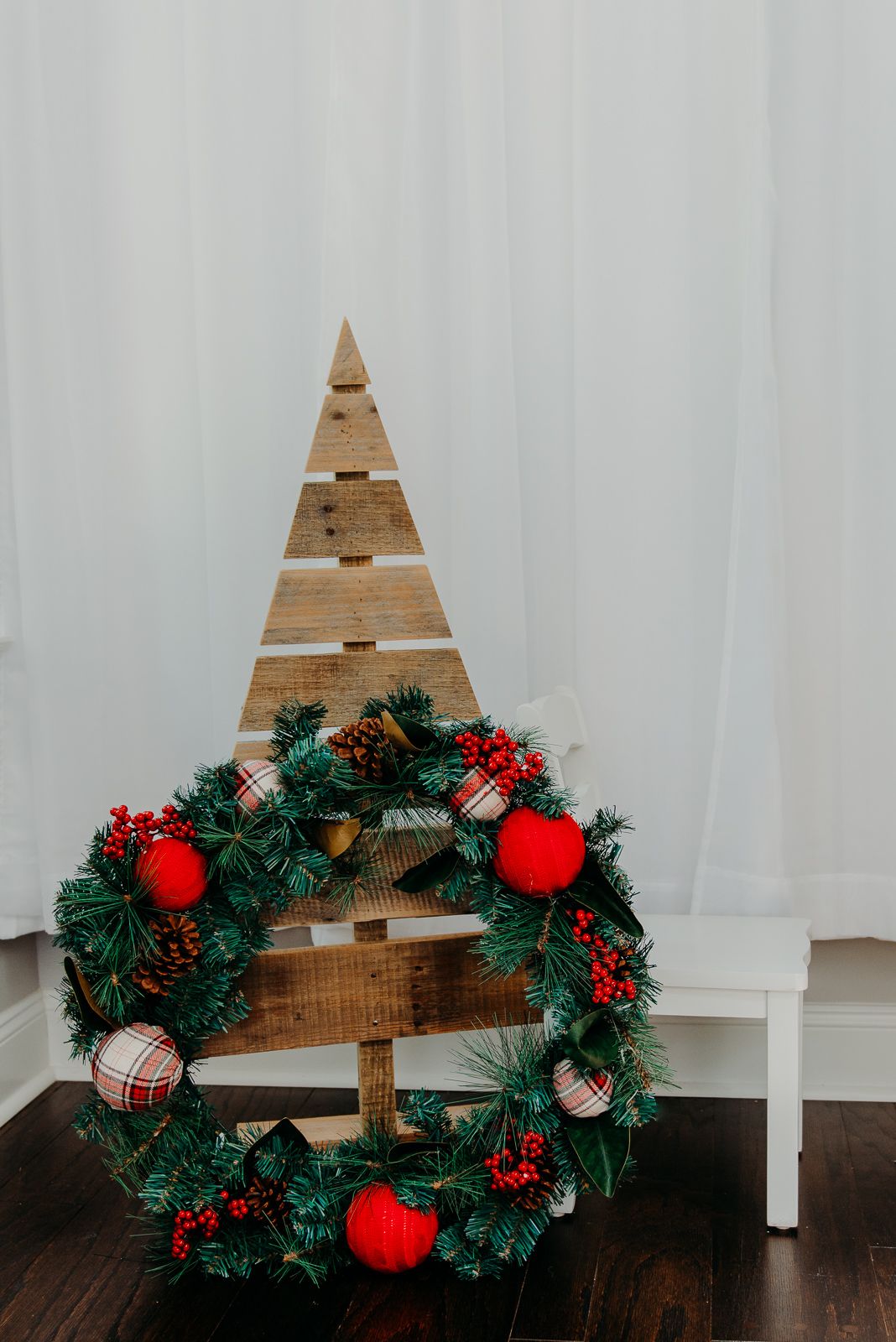 Wooden triangle tree display with traditional Christmas wreath decorated with red ornaments and pine cones.