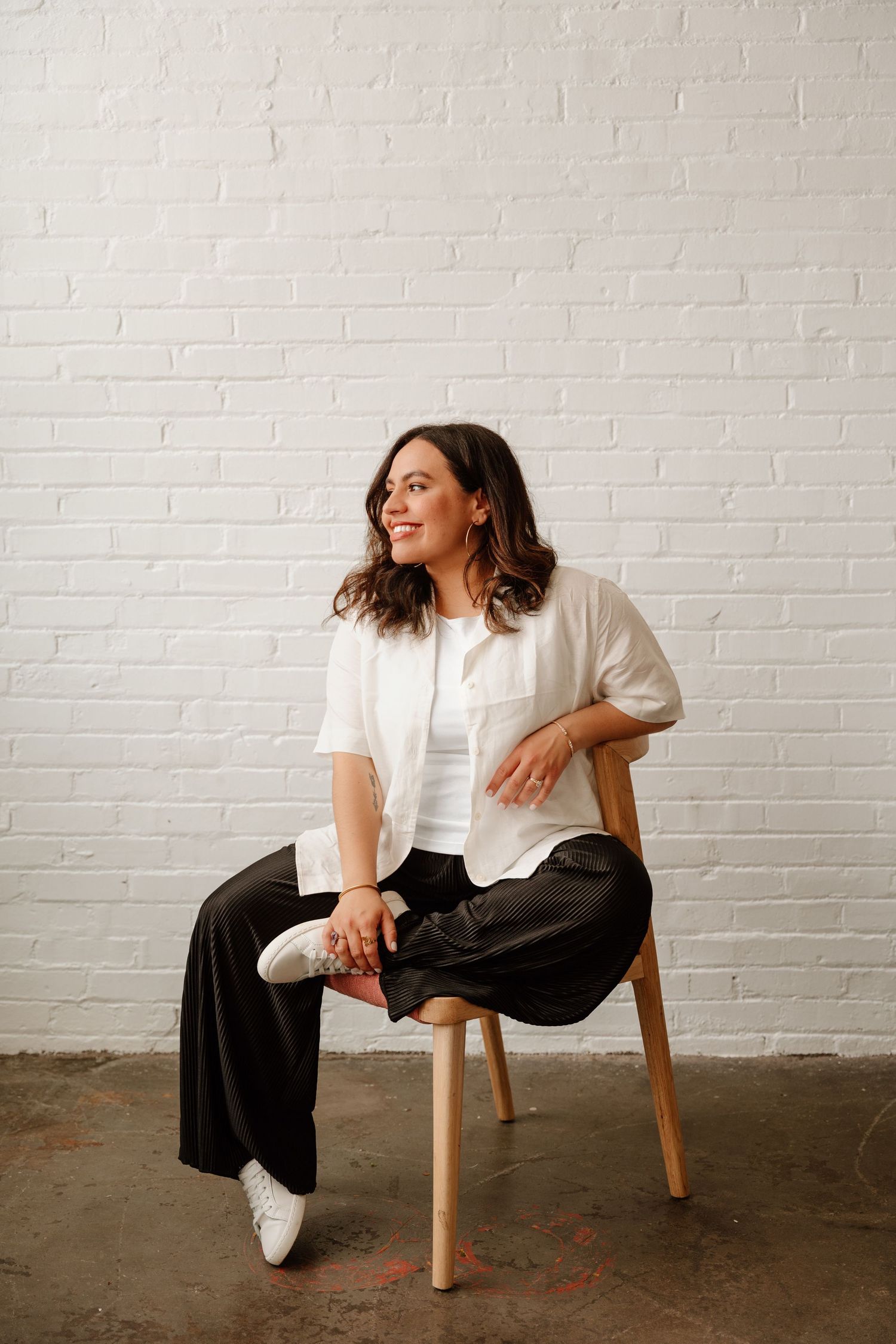 Person in white shirt and black pants sits casually on wooden chair against white brick wall in studio setting.