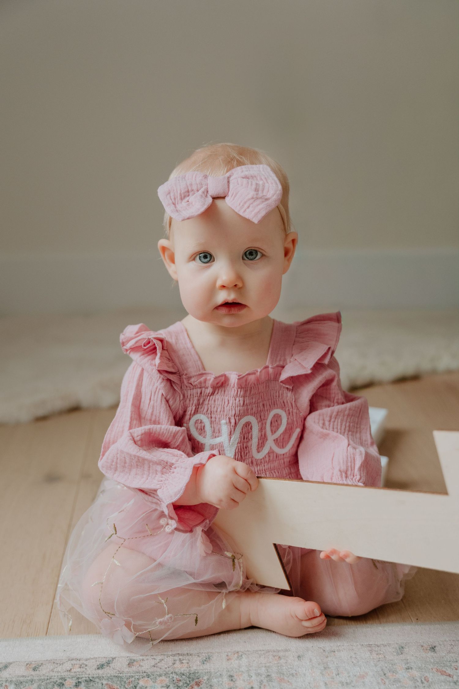 Baby wearing pink ruffled dress and bow sitting on floor with white decorative element.