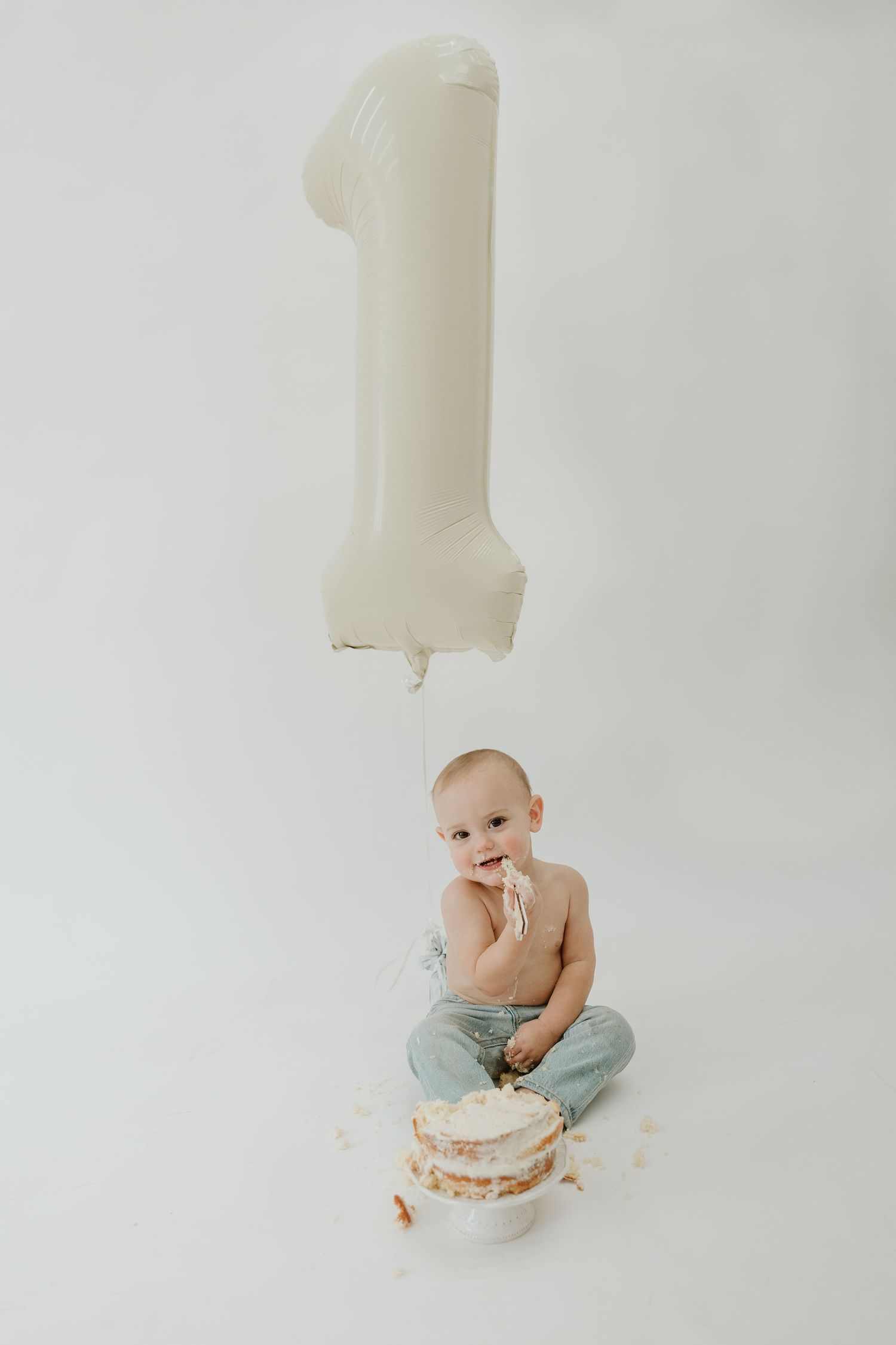 Baby in gray pants sits on white background while playing with cake smash dessert in artistic first birthday photo series.