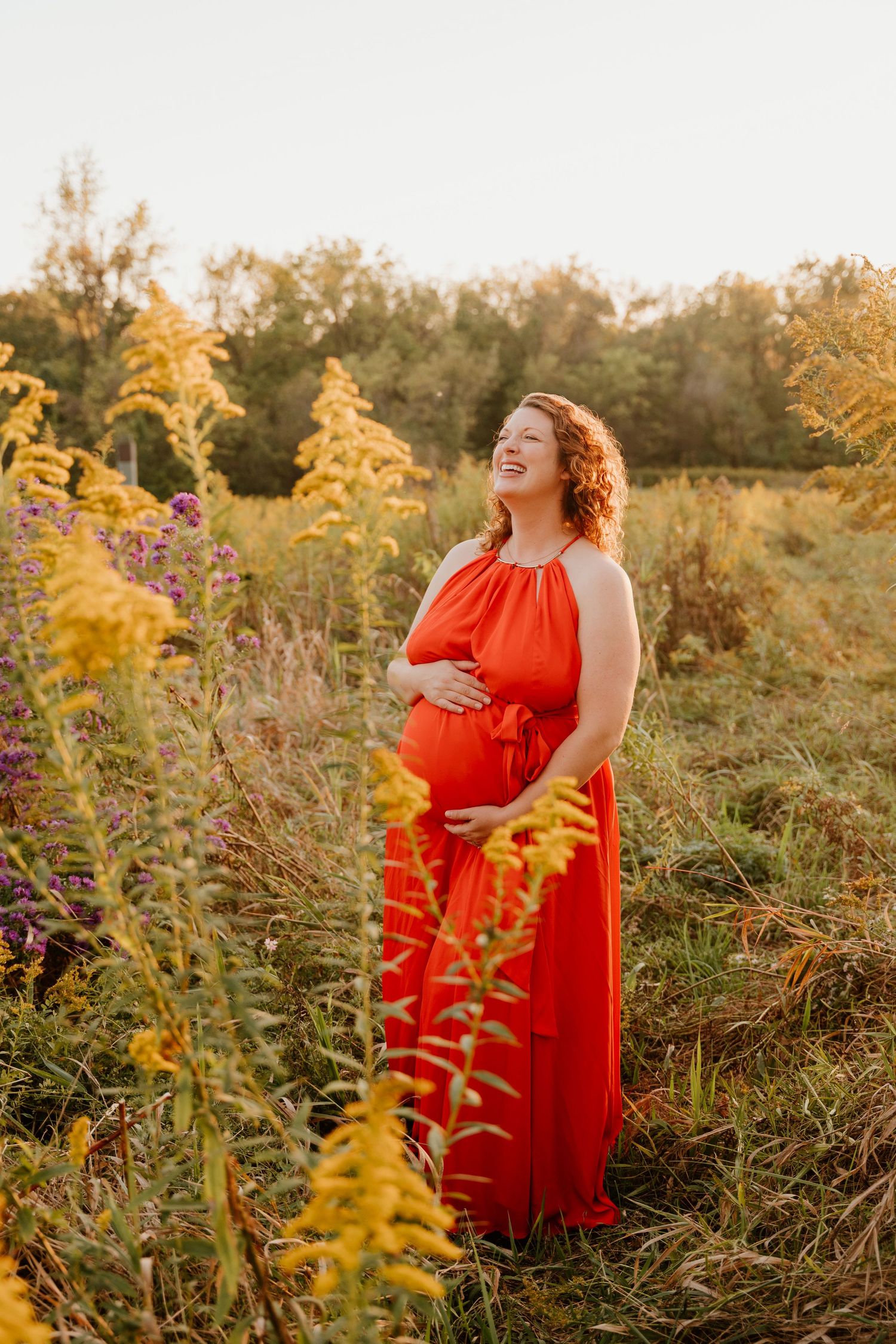 A person in a flowing red maxi dress stands joyfully among golden wildflowers at sunset in a natural field setting.