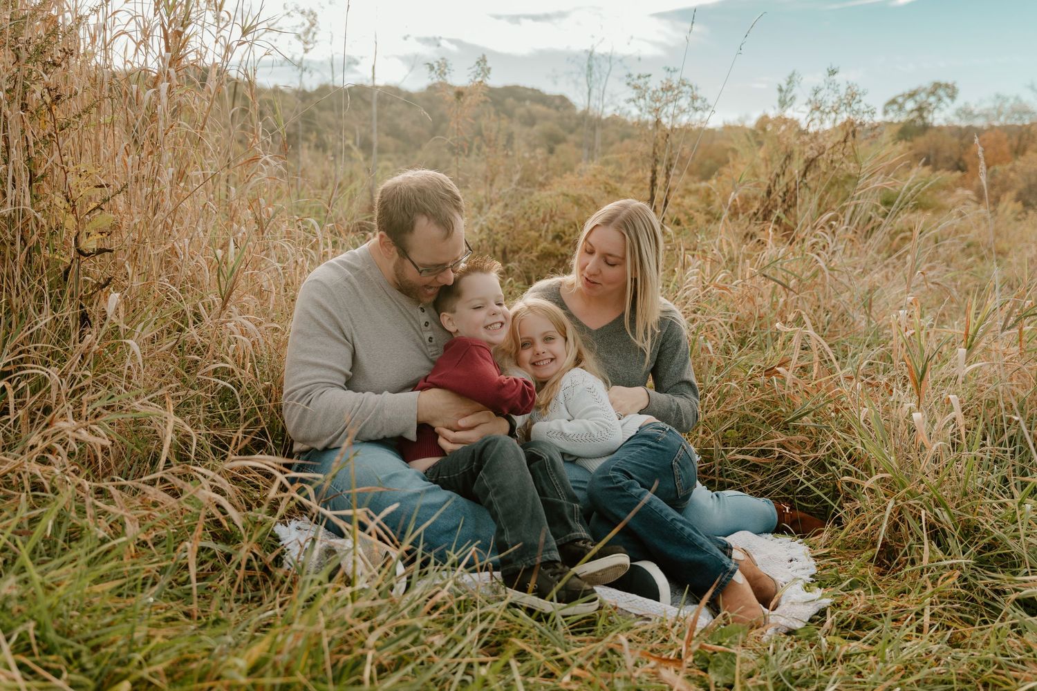 A family sits together on a blanket in tall autumn grass during a golden hour outdoor photo session.