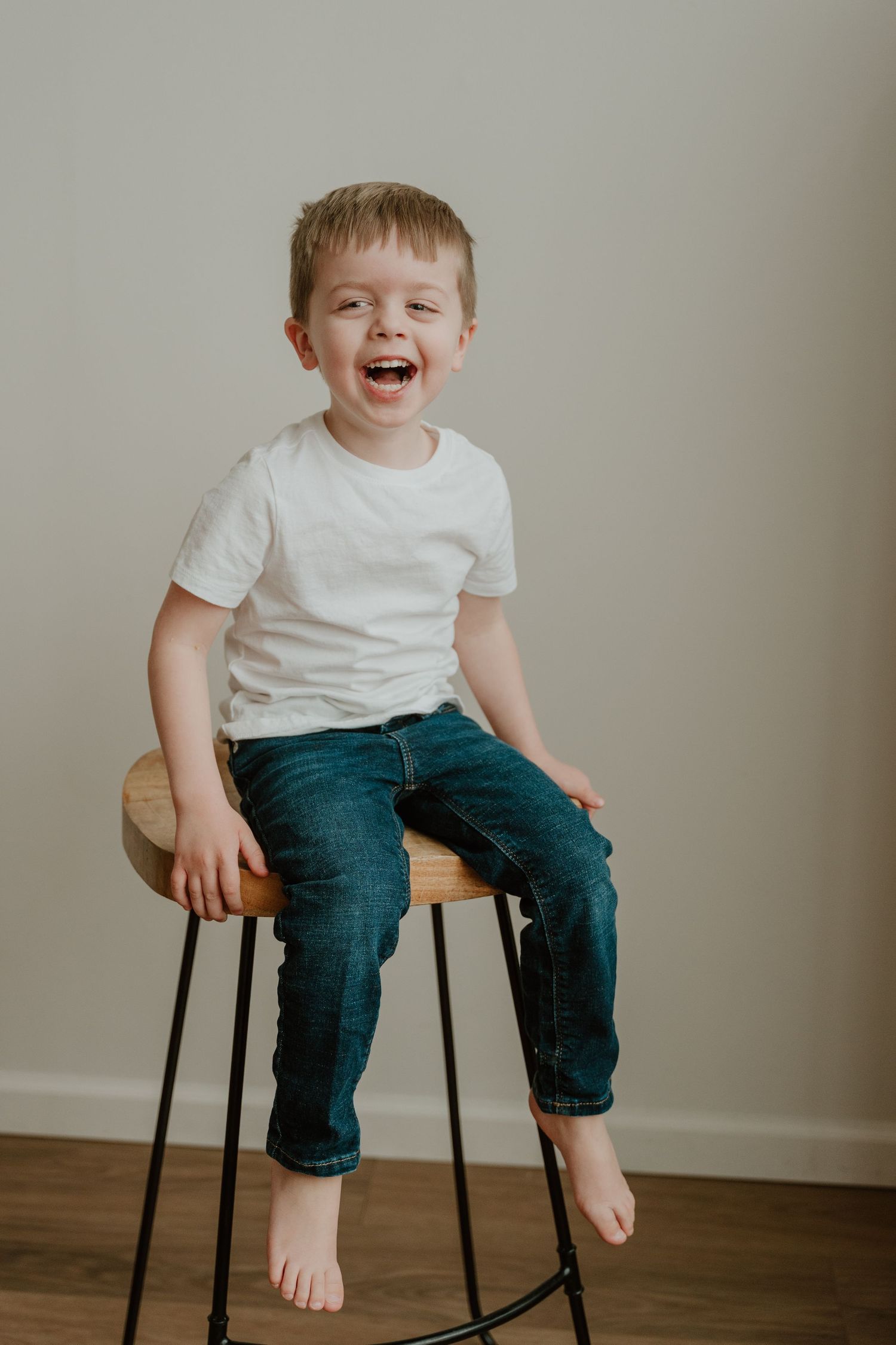 A small child wearing a white t-shirt and jeans sits barefoot on a dark stool, laughing joyfully against a neutral background.