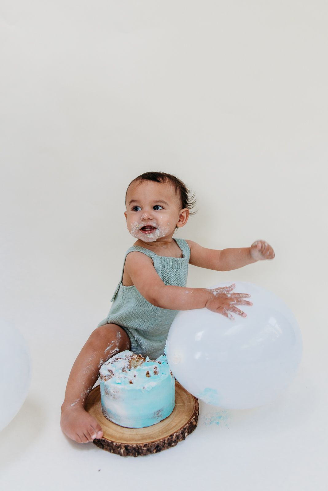 Smiling baby in turquoise outfit with birthday cake and white balloons.