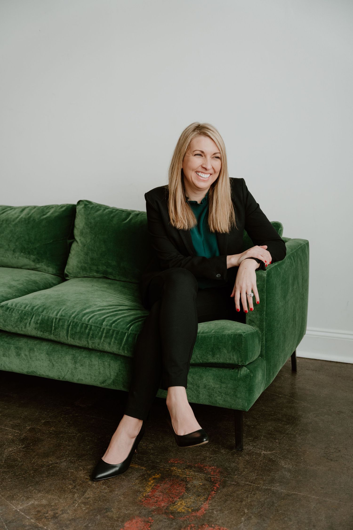 Professional looking woman sitting casually on a plush emerald green velvet sofa against a light gray wall.