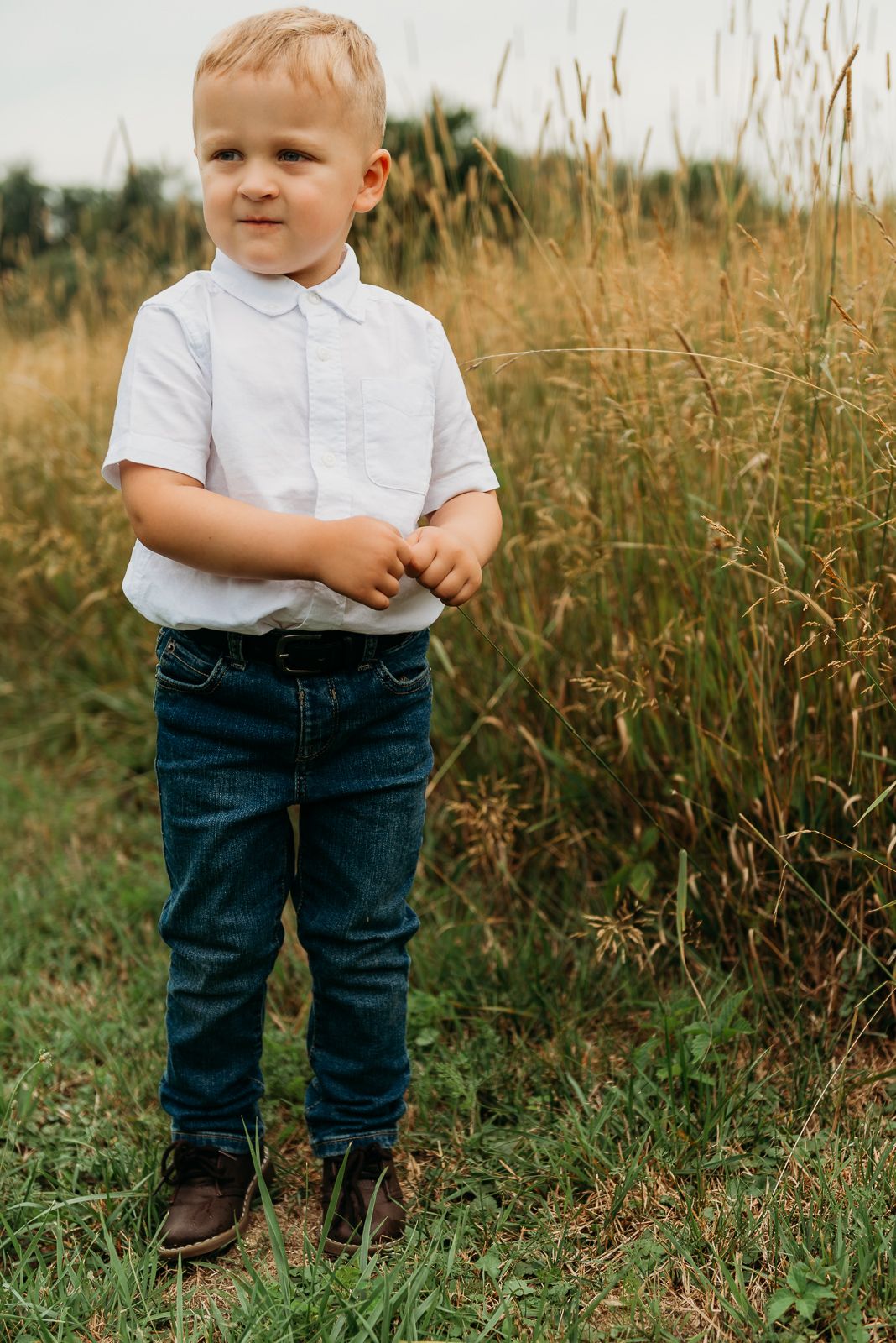 Young child in white polo shirt and jeans standing in tall grass field.