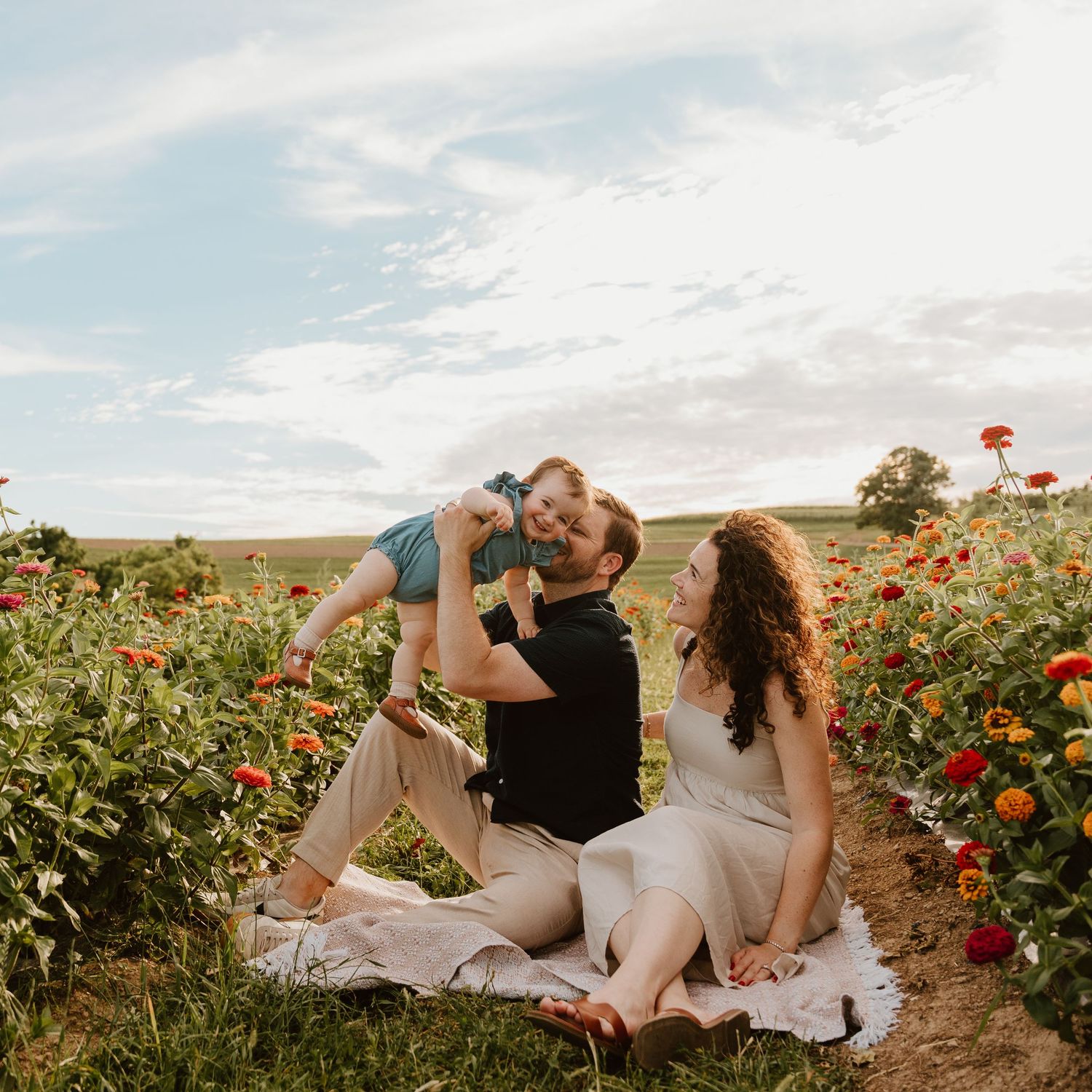 A joyful family sits in a vibrant flower field, with a man lifting a baby while a woman smiles beside them.