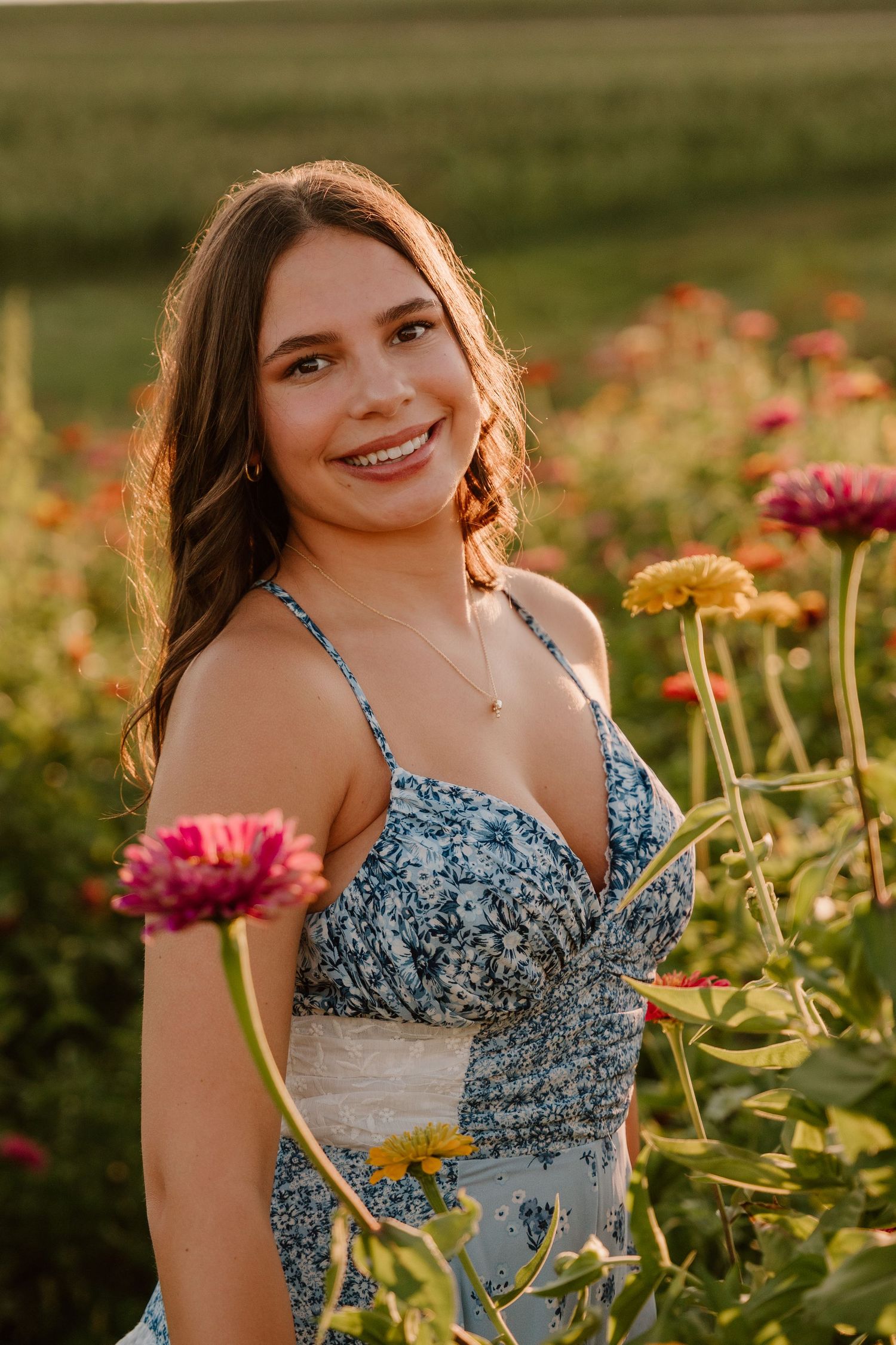 Young woman in floral sundress smiling among colorful zinnia flowers in a garden at golden hour.