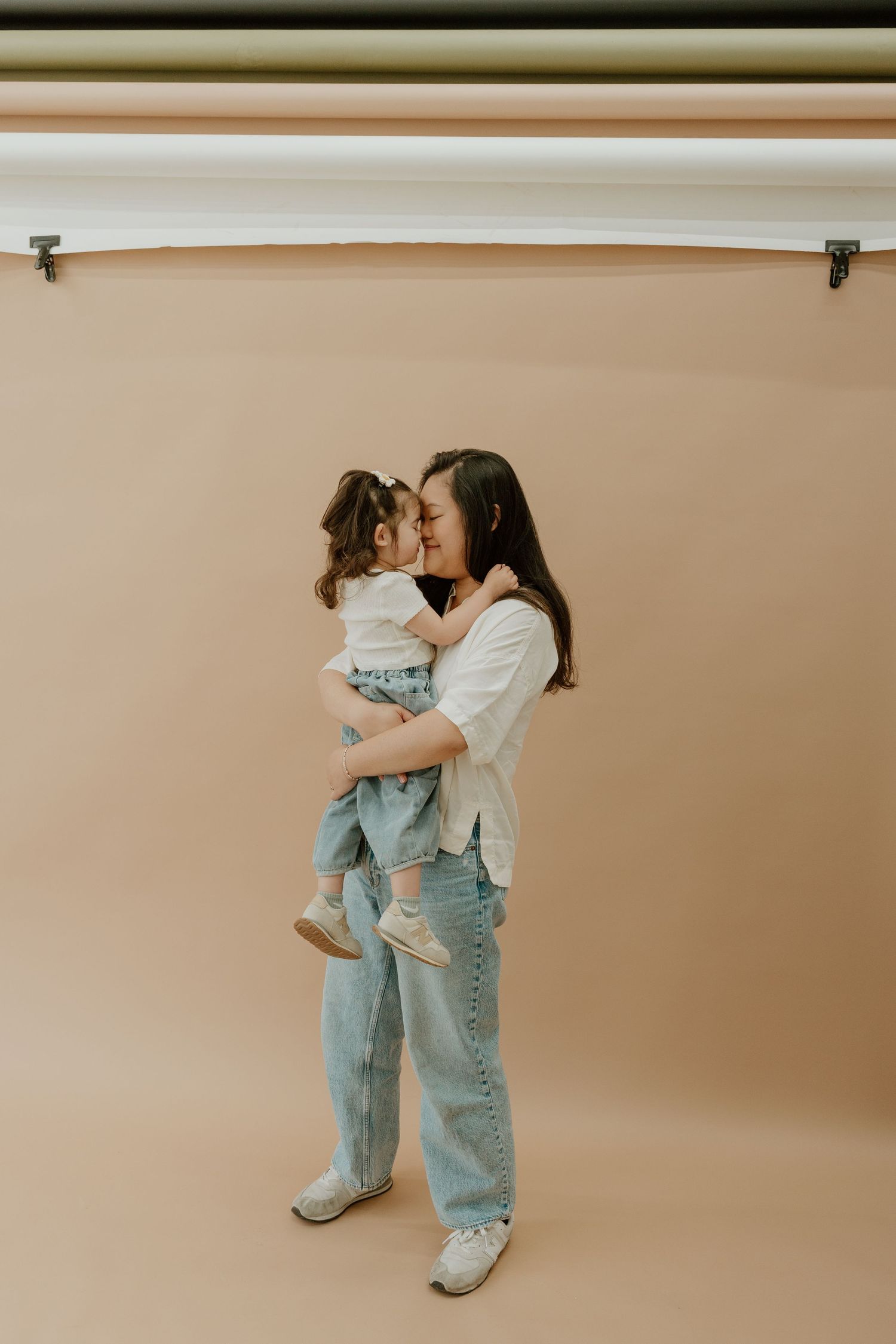 Portrait of parent and child embracing in casual outfits against beige backdrop in studio setting.