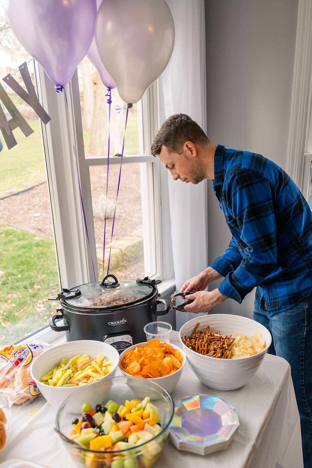 Party buffet table setup with colorful balloons, featuring various dishes and a person in blue plaid serving food.