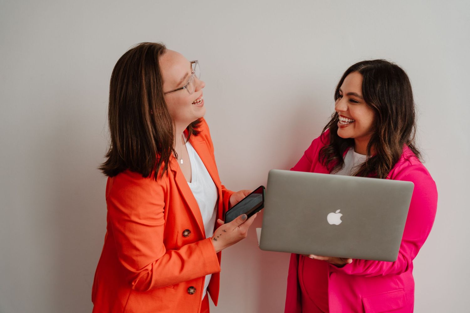 Two colleagues in bright blazers share a laugh while looking at a MacBook laptop against a white wall.