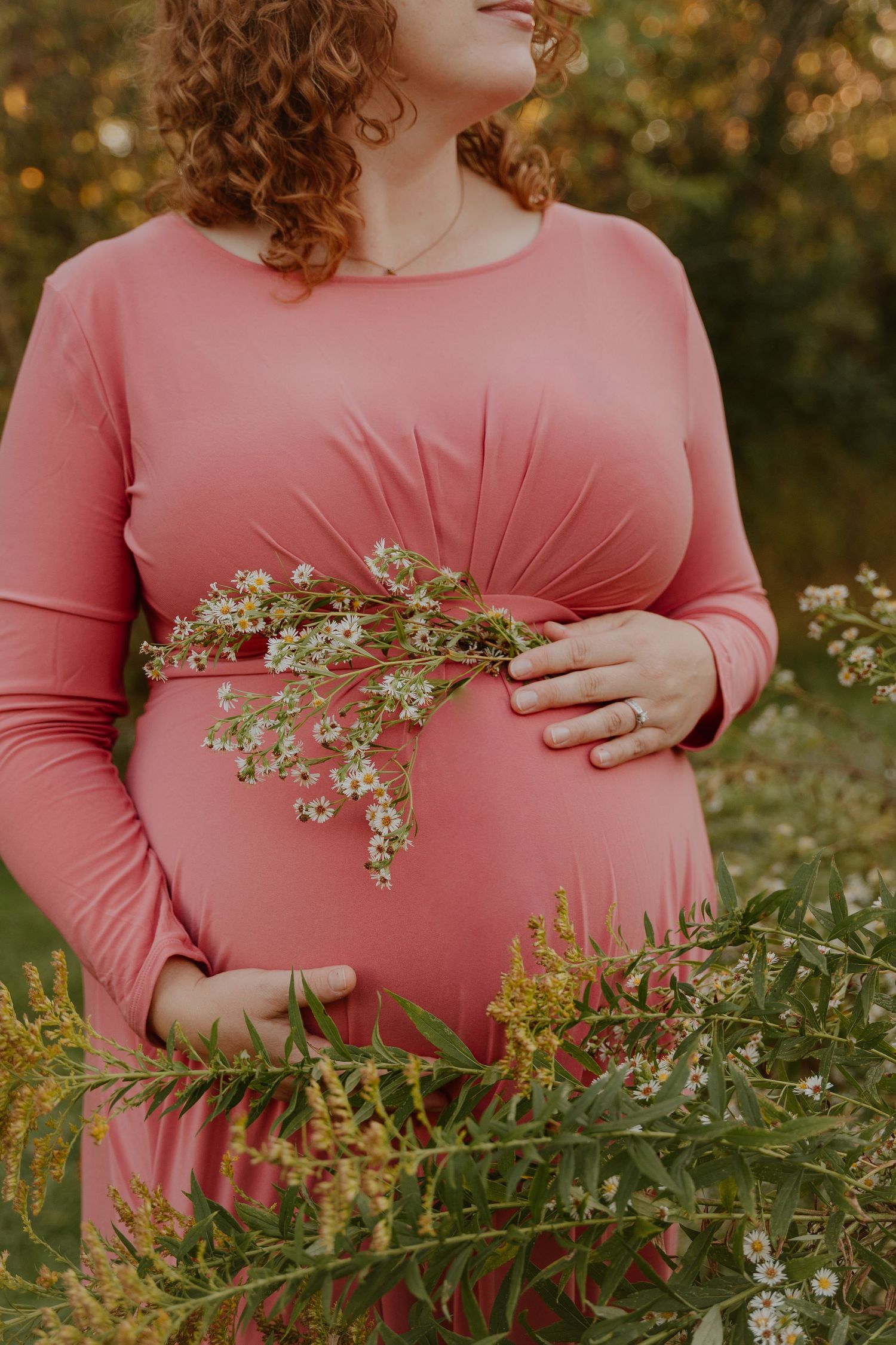 A person in a pink maternity dress holds delicate white wildflowers against their pregnant belly in a natural outdoor setting.