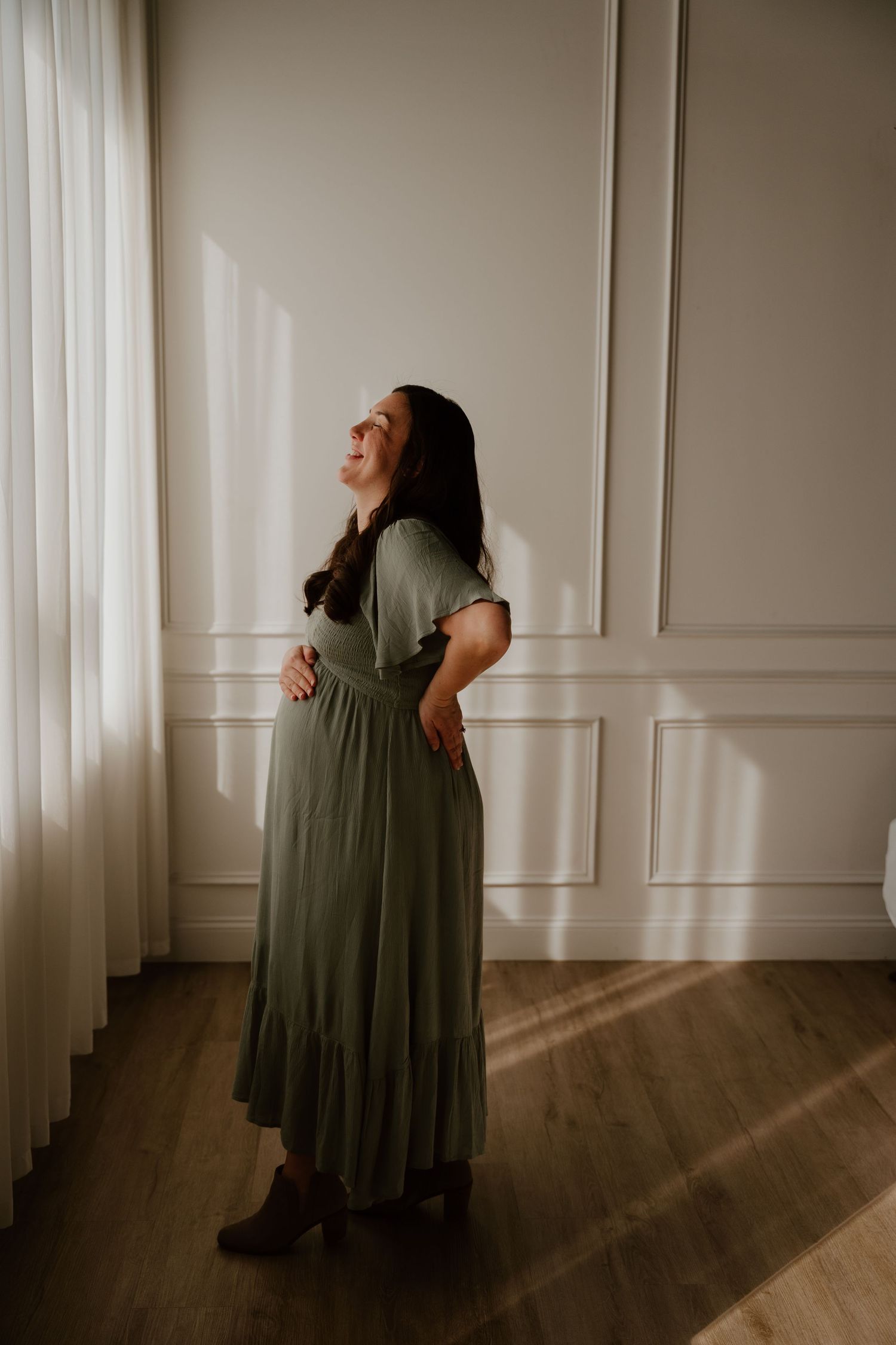 A person in a flowing sage green maxi dress stands by a window, casting dramatic shadows on hardwood floors.