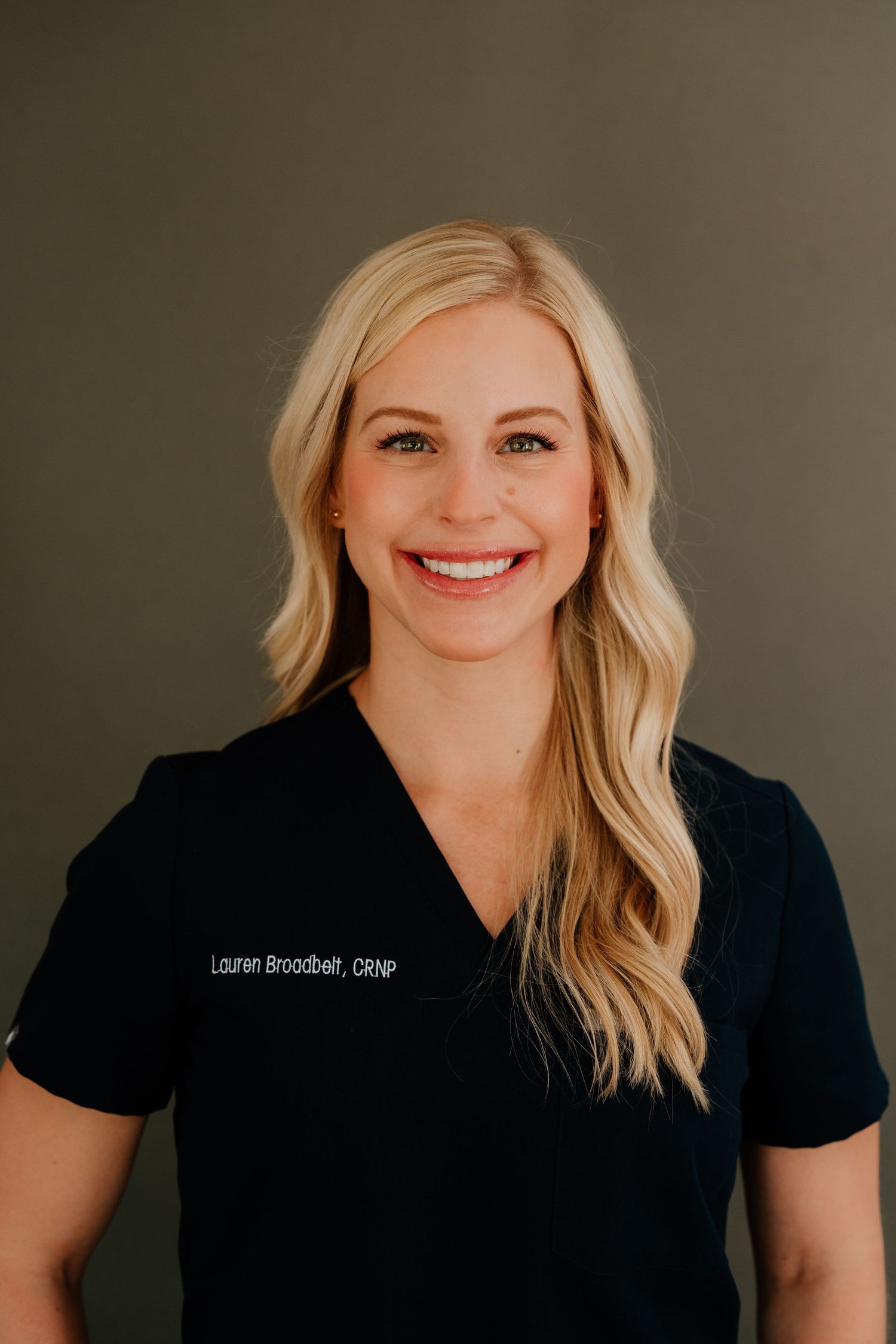 Healthcare professional in black scrubs with blonde wavy hair smiling warmly against neutral background.