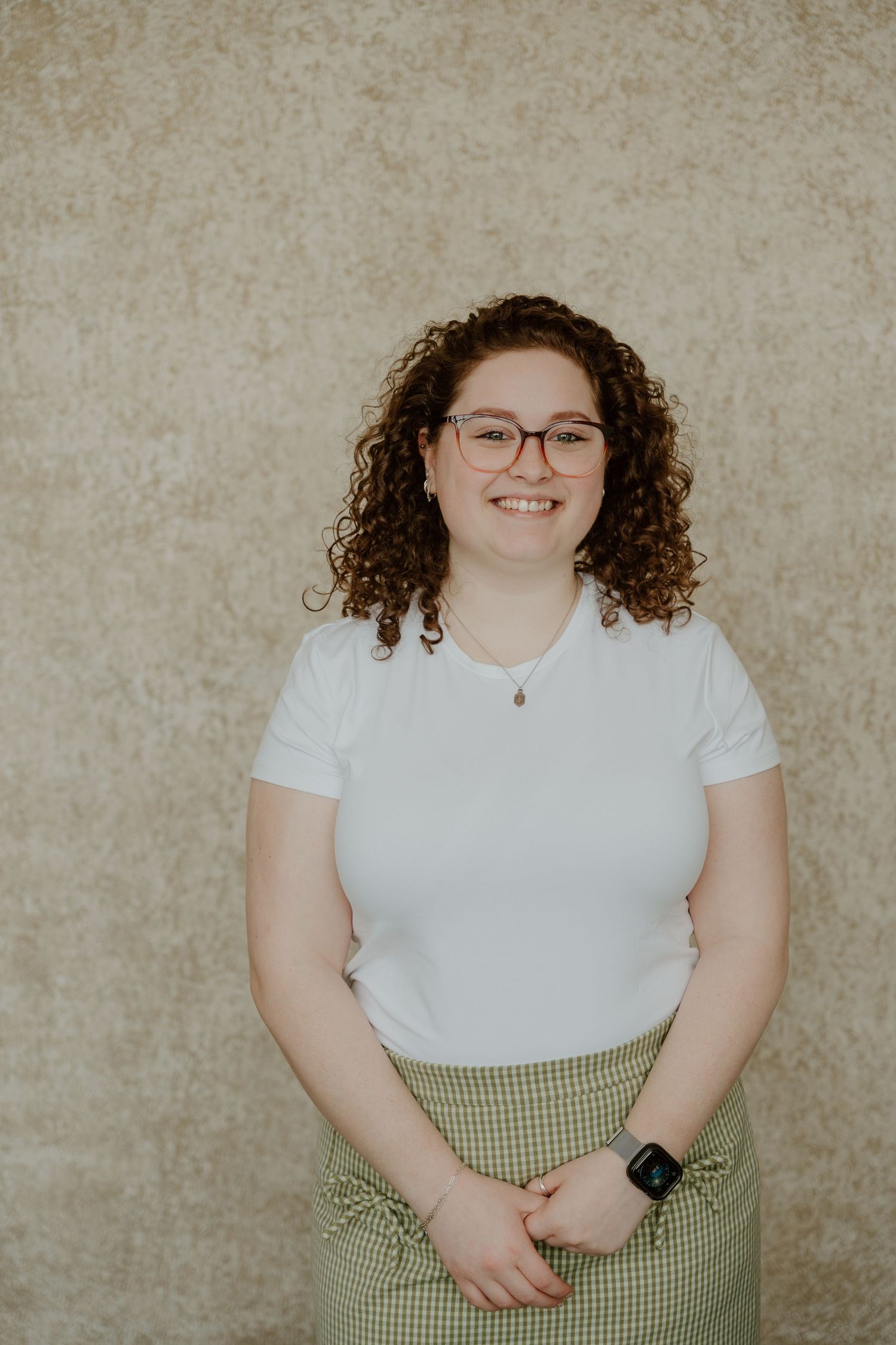 Professional portrait against beige background showing person in white top and gingham skirt with curly hair and glasses.