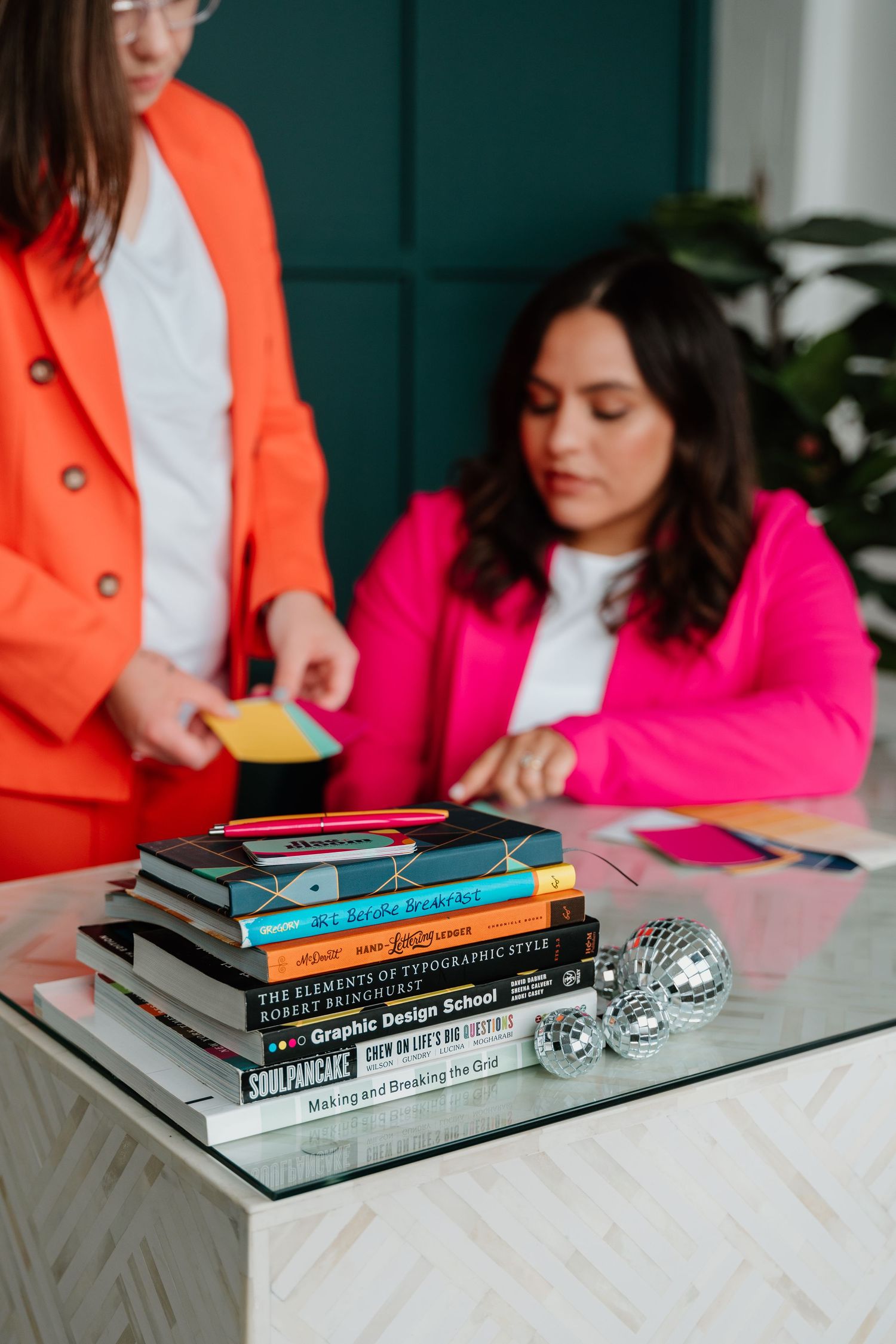 A collection of books arranged on a marble table with people in orange and pink attire studying in the background.