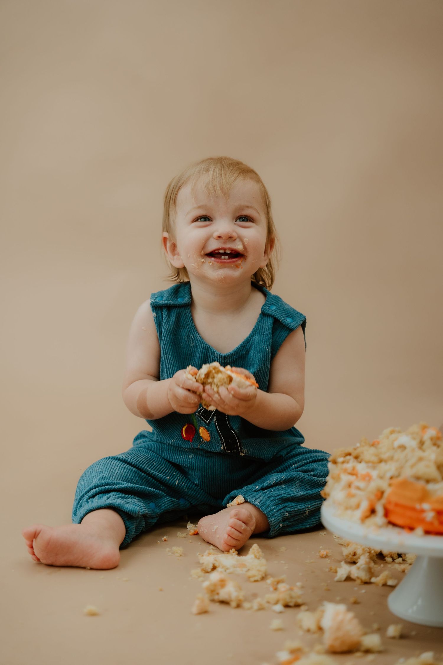 A series of photos showing a baby in teal overalls having fun during a cake smash photo session against a beige backdrop.