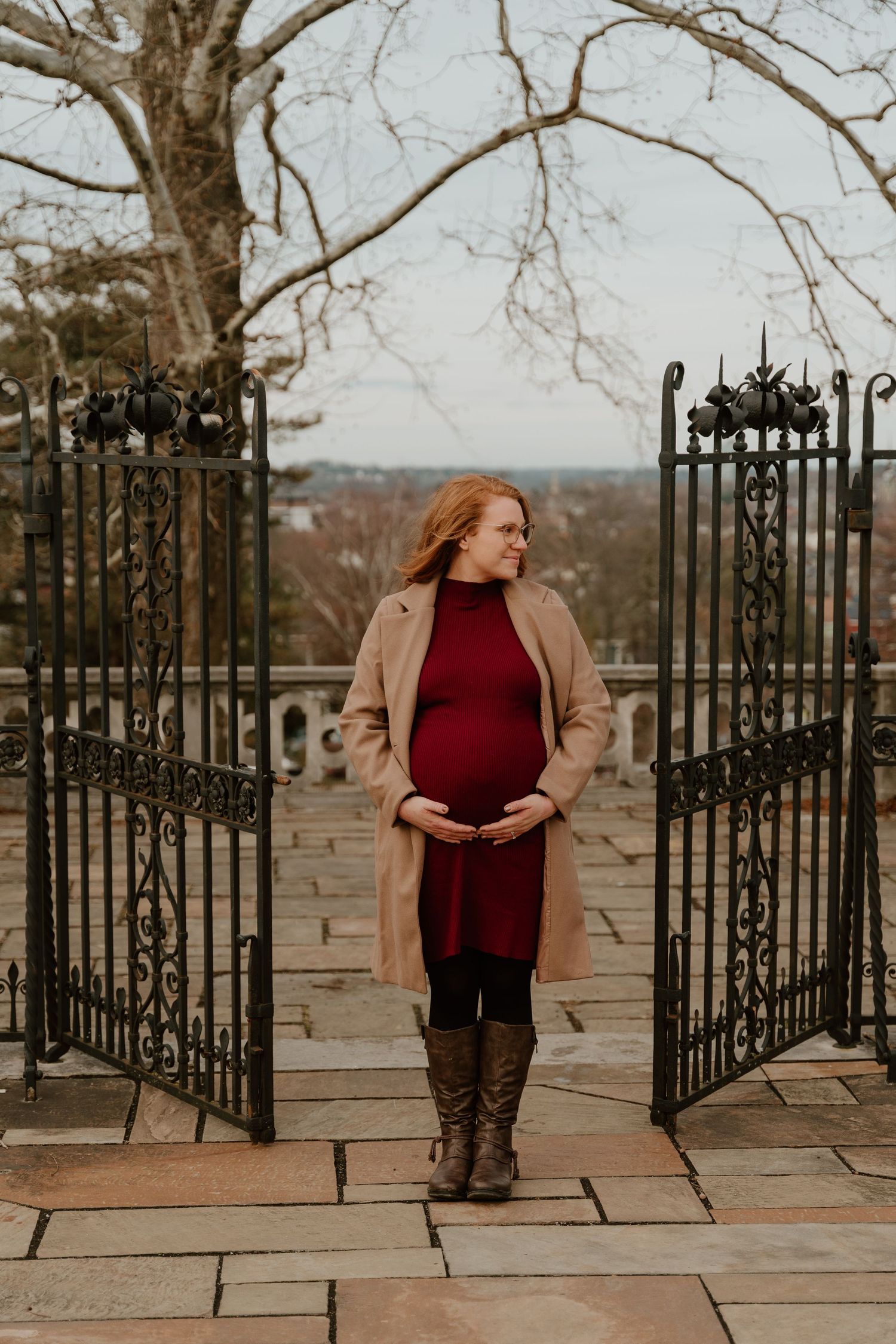 Expectant mother in a burgundy dress and tan coat stands between ornate iron gates on a stone terrace during autumn.