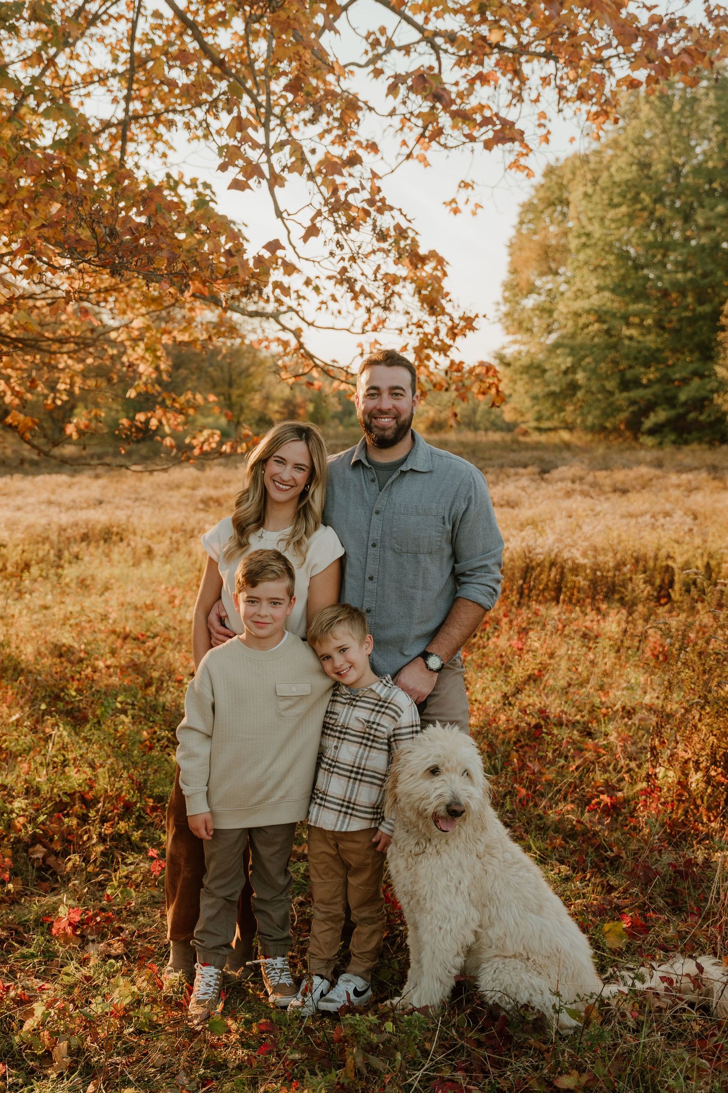Family and white fluffy dog pose together in autumn field during golden hour sunset.