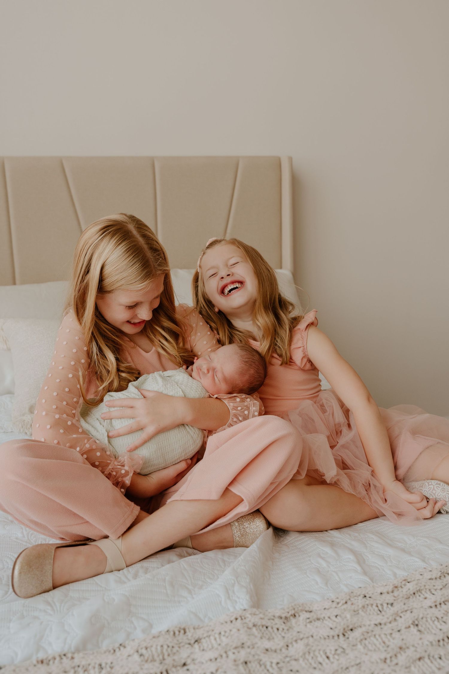 Three siblings share a joyful moment together on a bed with neutral decor and soft pink accents.