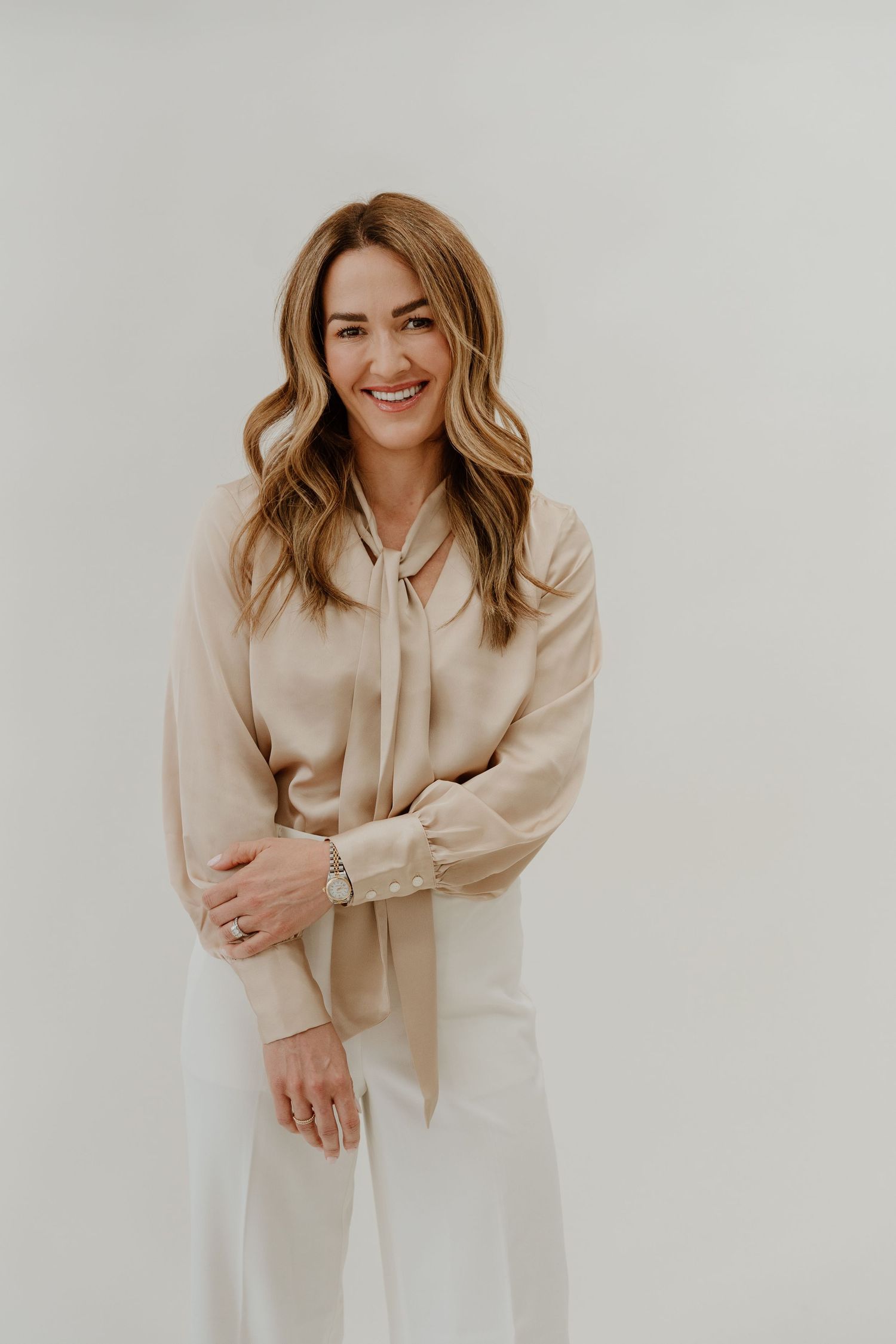 Professional studio portrait of someone in a beige silk blouse and white pants standing against a light background.