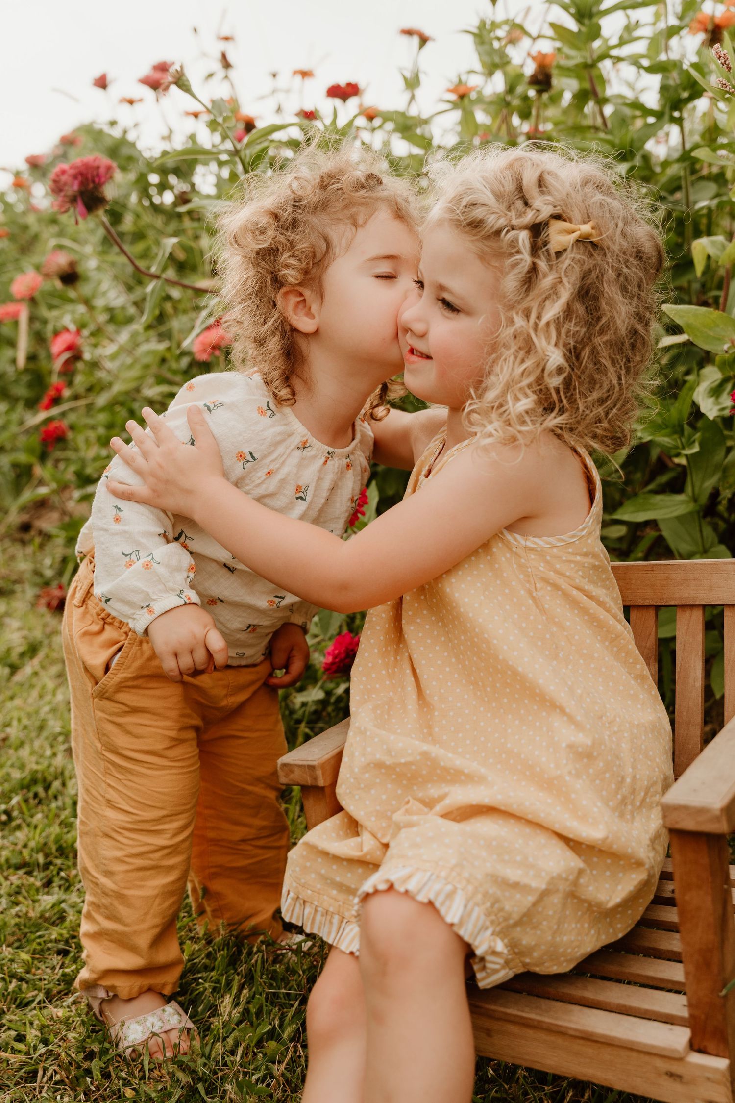 Two curly-haired toddlers share a sweet kiss on a wooden bench surrounded by colorful blooming flowers outdoors.