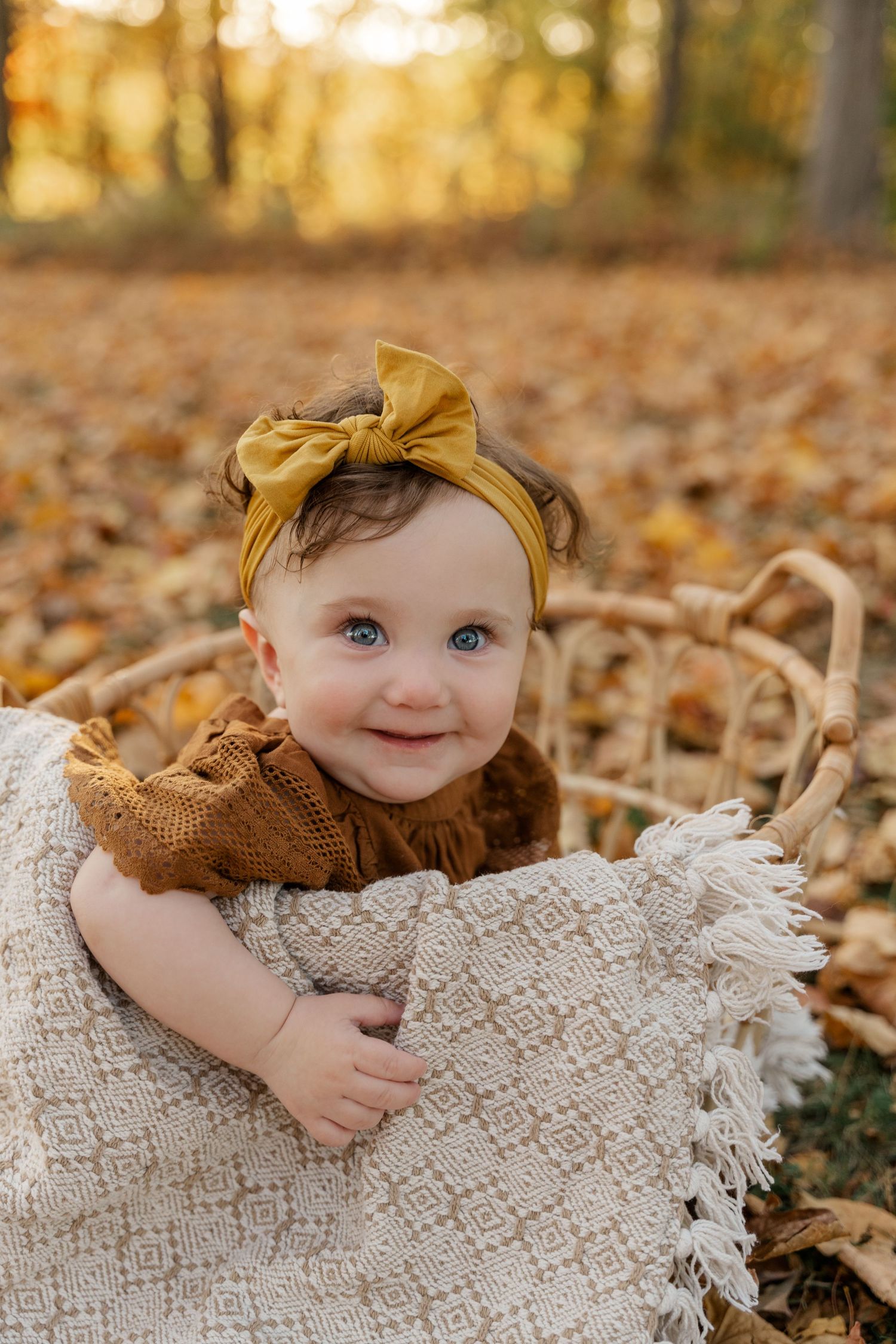 Baby in yellow headband and brown sweater on textured blanket with autumn leaves.