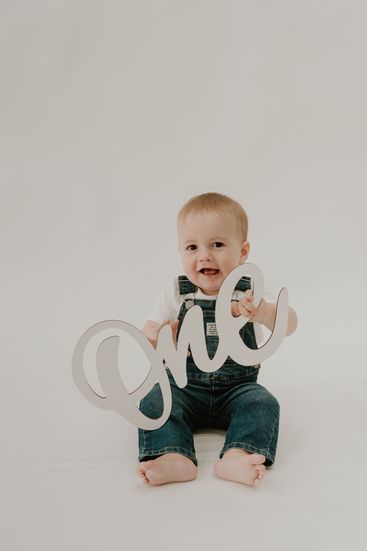 A baby in denim overalls sits on a white background in a series of milestone photos with a metallic 'one' sign.
