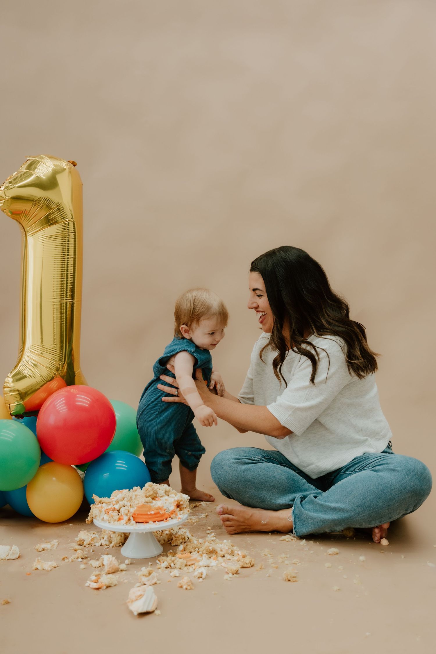 First birthday celebration with colorful balloons, cake smash setup, and joyful interaction on beige backdrop.