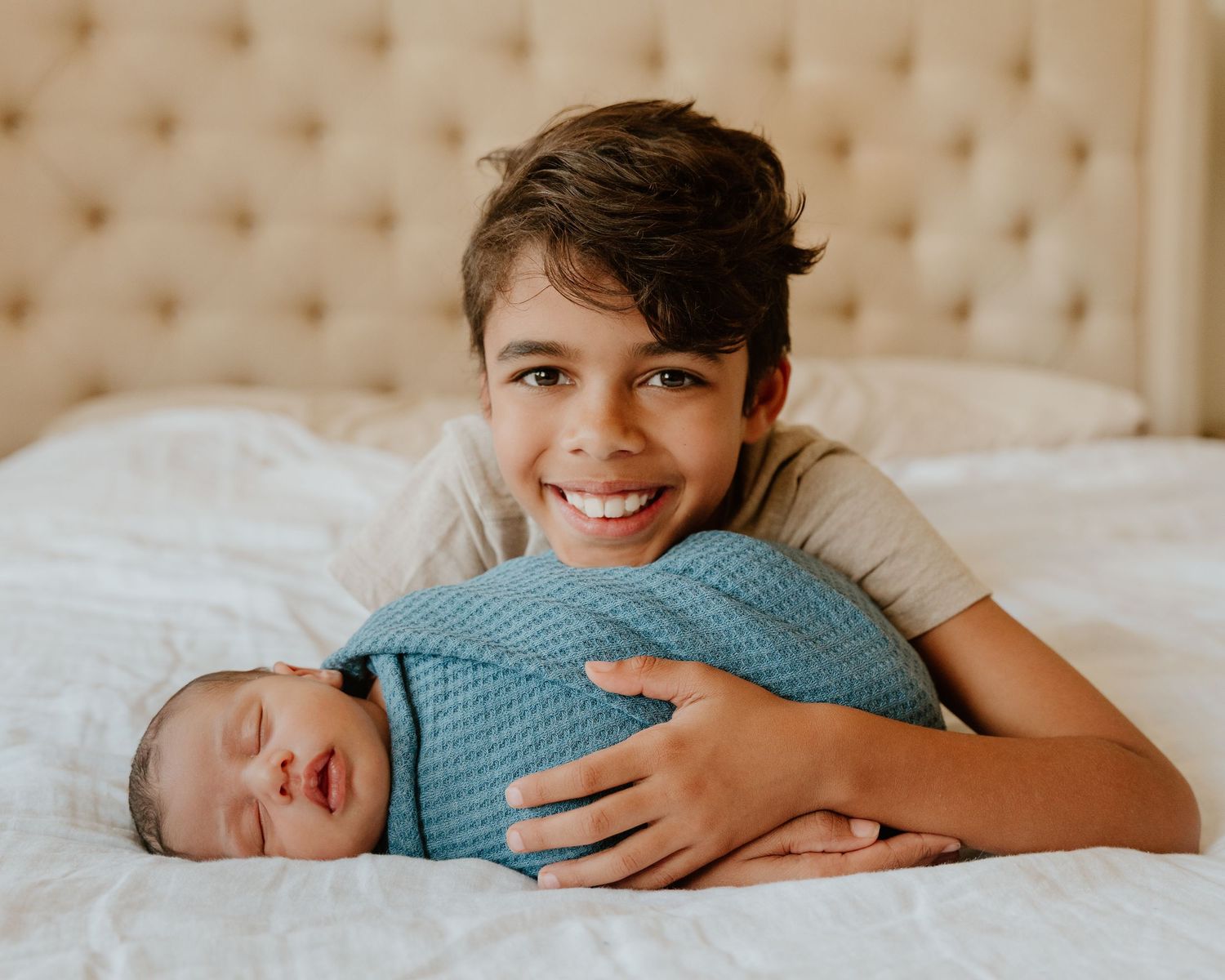 Two children lie side by side on white bedding while wearing matching blue shirts and sharing a tender moment.