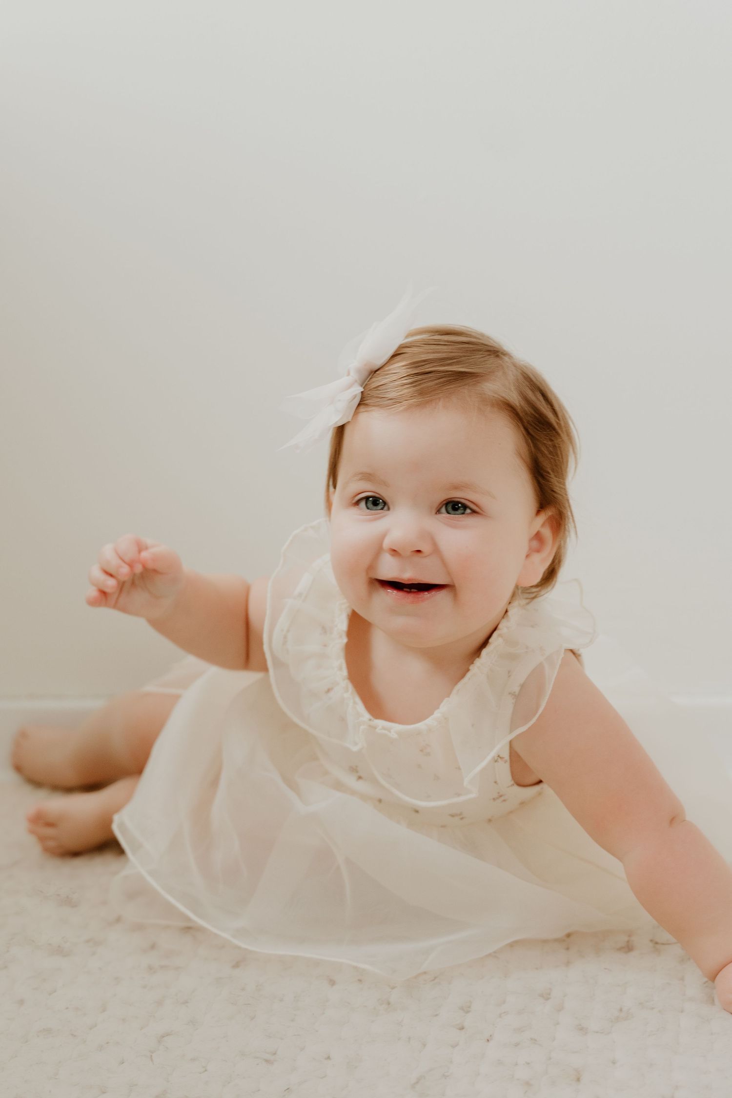 A baby in a white ruffled dress crawls and smiles on a light colored carpet.
