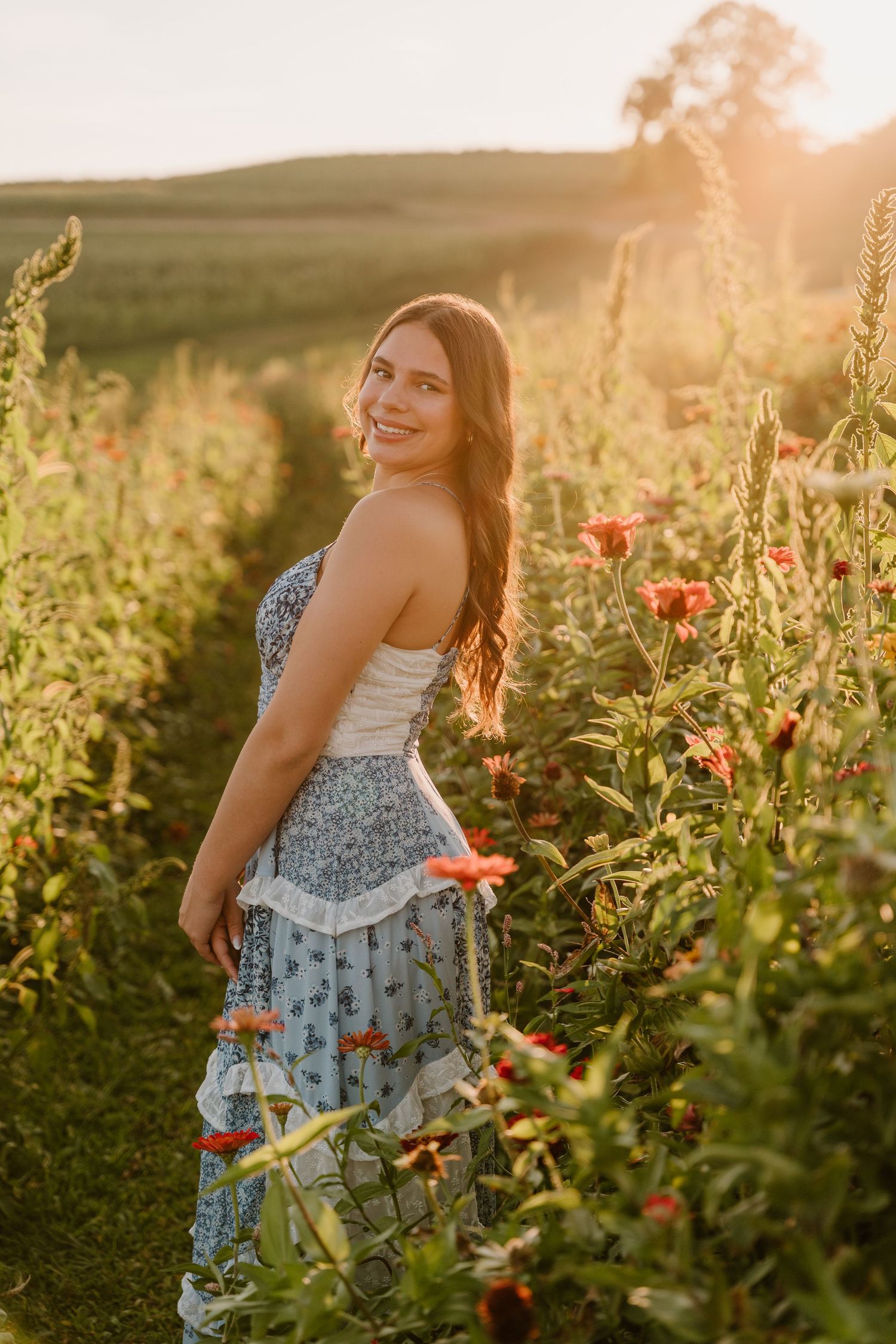 A person in a blue floral dress stands joyfully among blooming wildflowers during golden hour sunset.