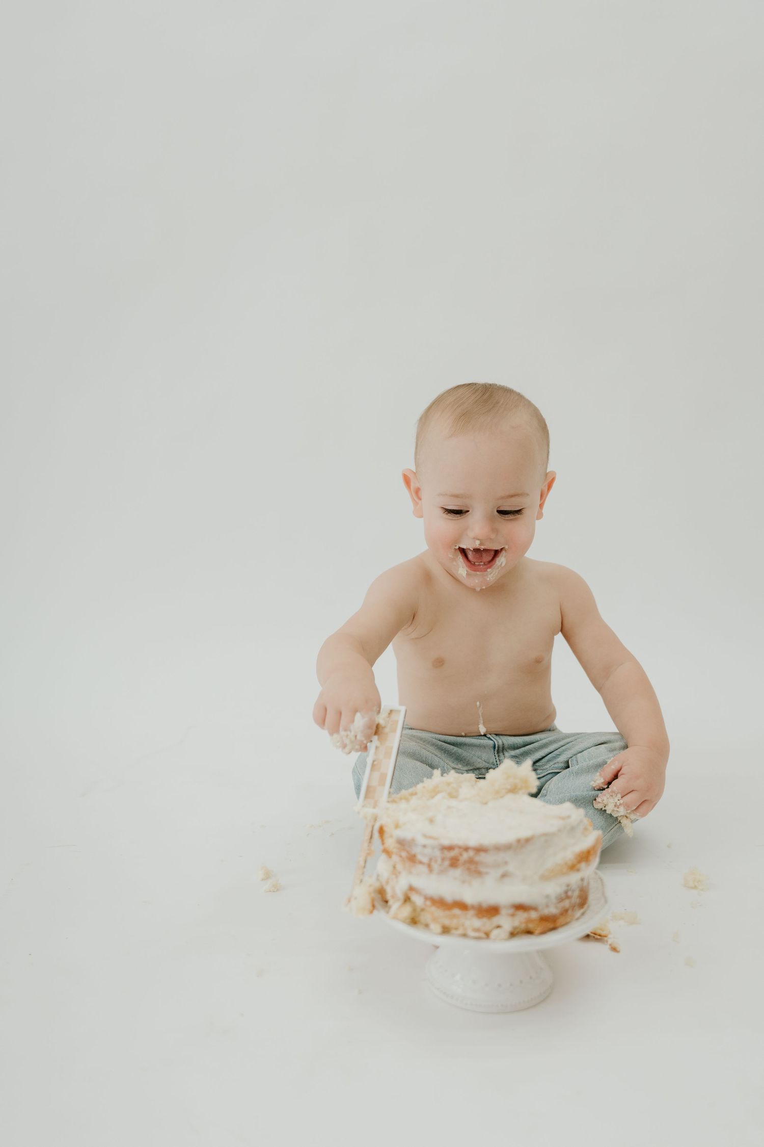 A joyful baby sits and plays with a white frosted naked cake on a plain white background.