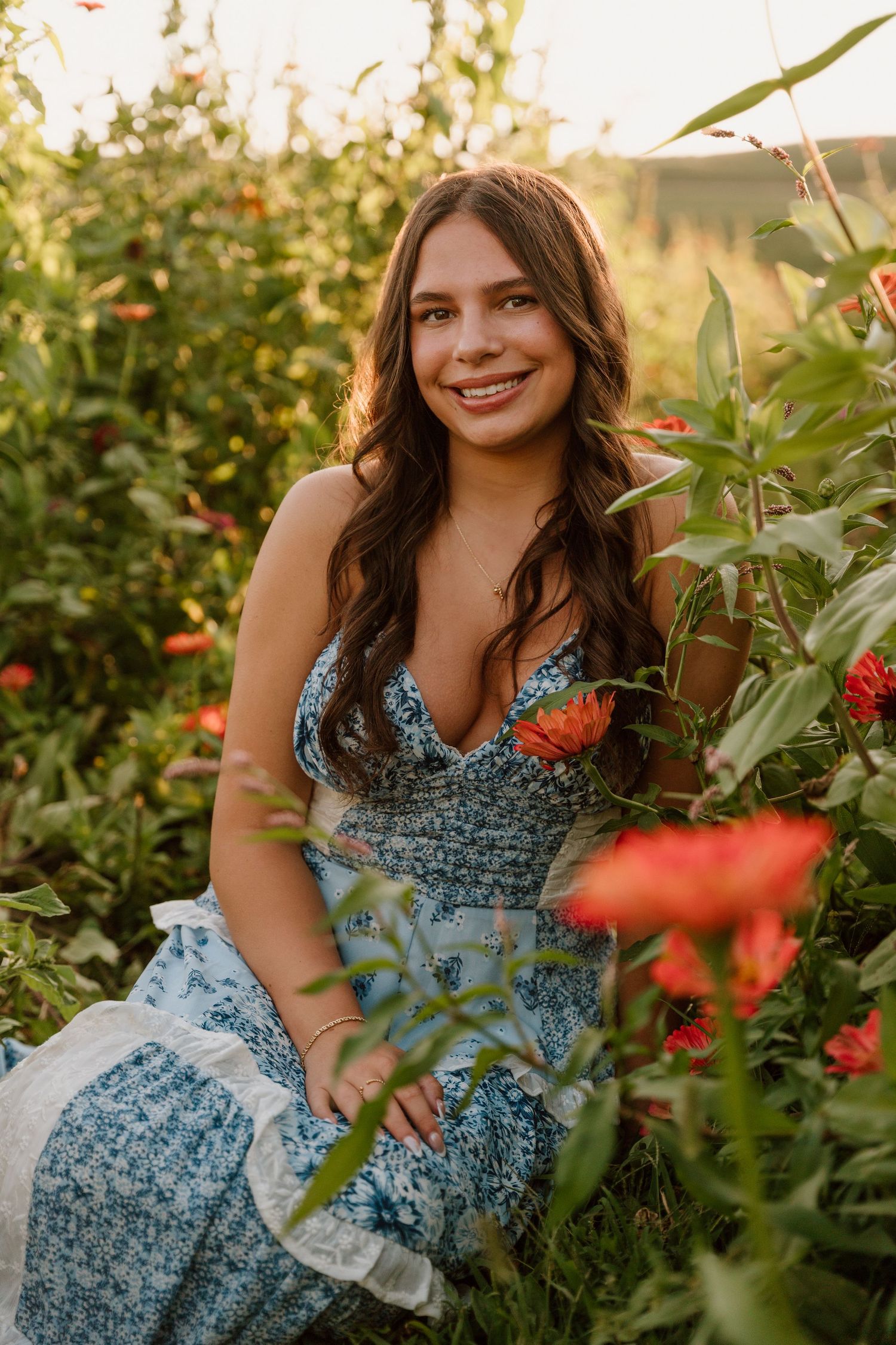 Smiling young woman in blue floral maxi dress sitting among red flowers in a sunlit garden.