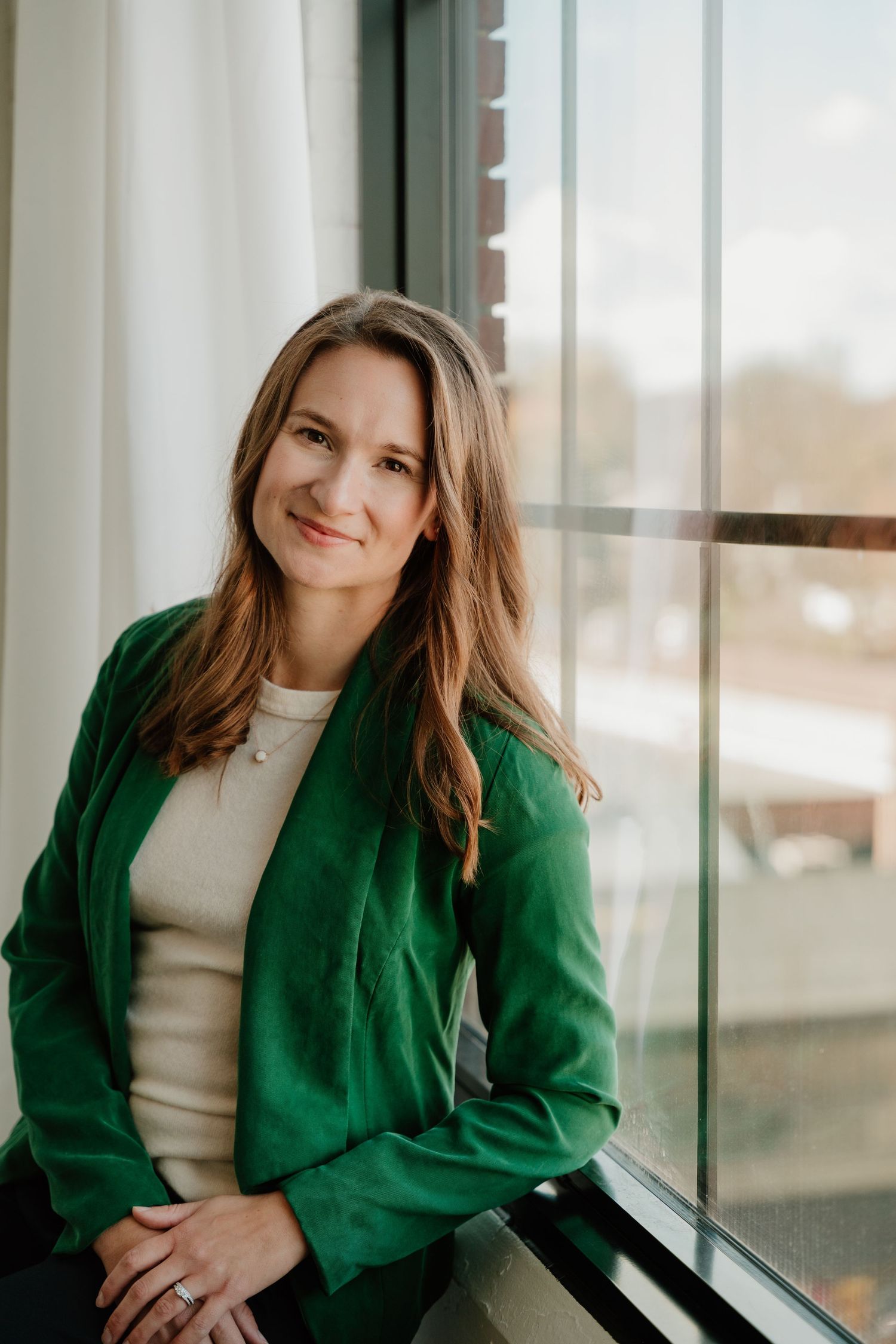 Professional portrait by a window showing someone in a bright emerald green blazer and cream top with natural lighting.
