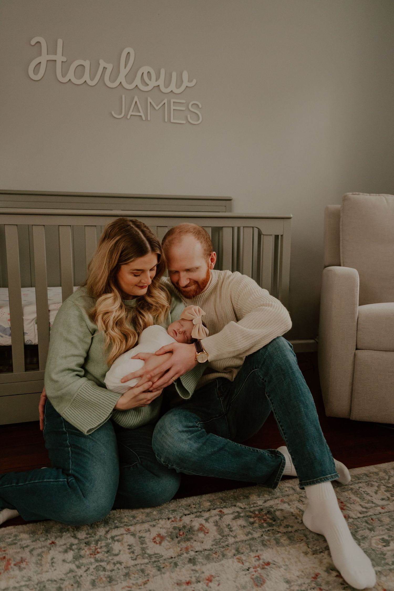 A couple sits on the floor of a nursery with their newborn baby beneath wall text reading 'Harlow James'.