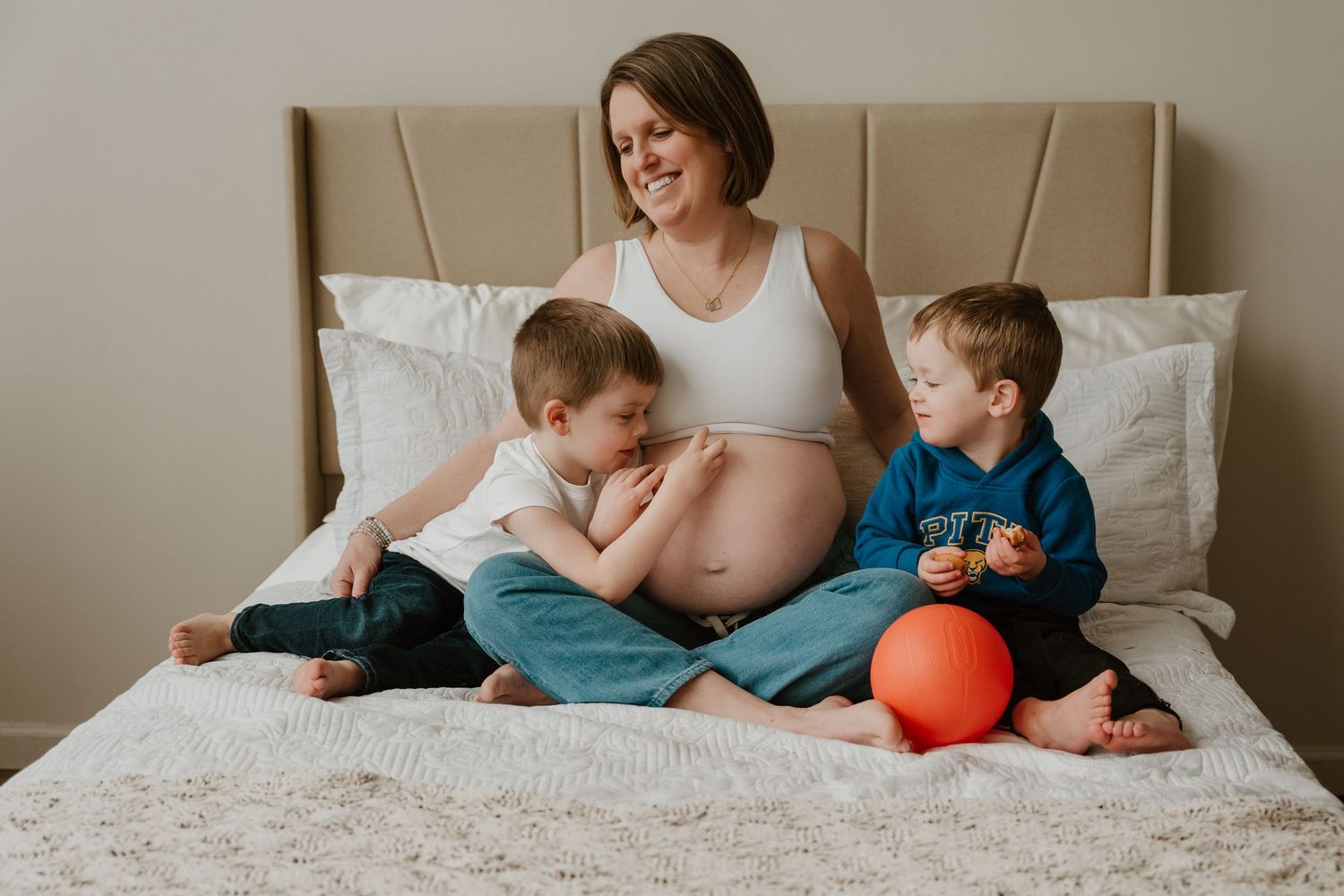 Pregnant woman in white tank top relaxes on bed with two young children and orange ball.