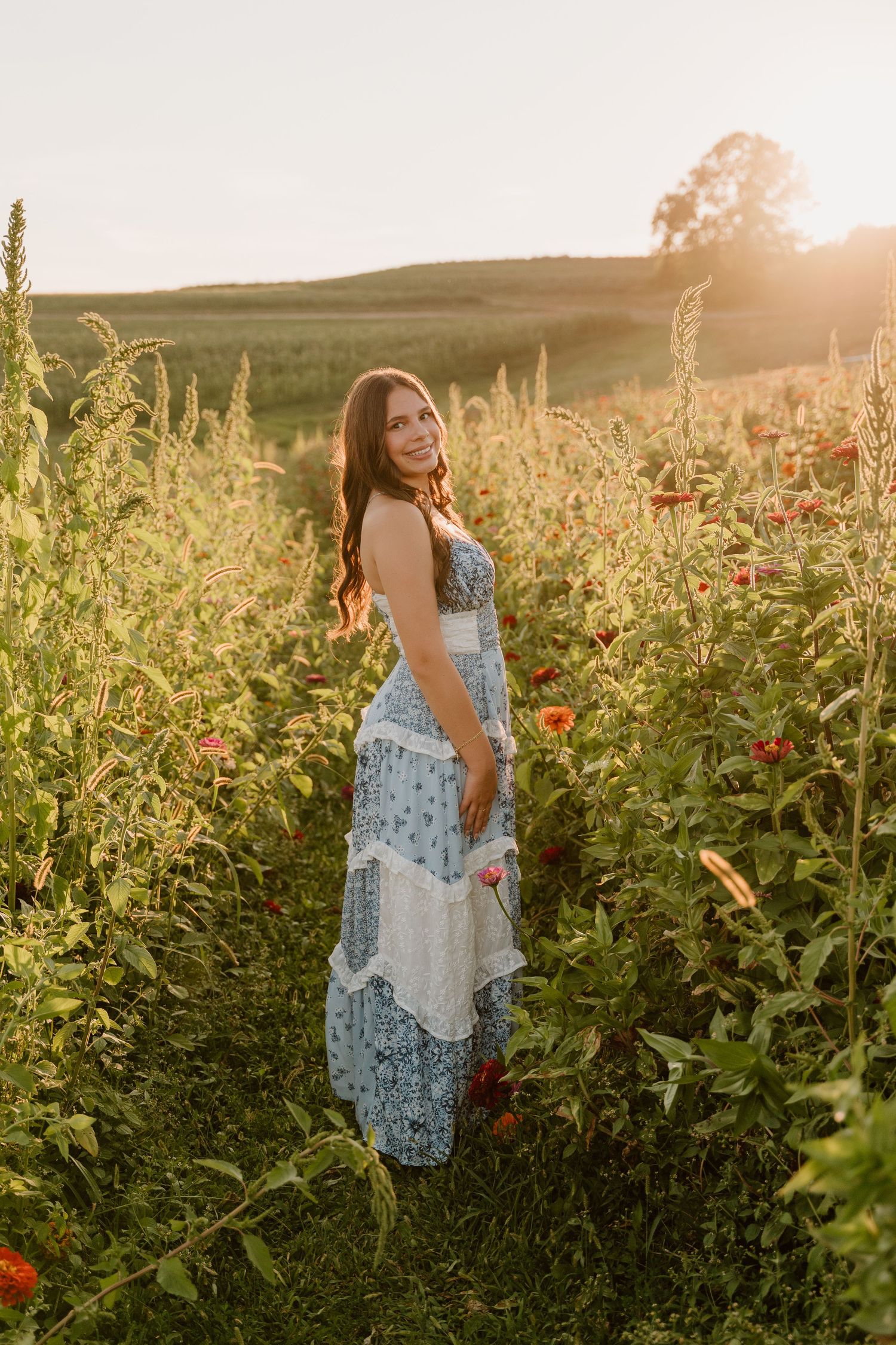 Smiling young woman in blue patchwork maxi dress standing in a vibrant flower field during golden sunset.