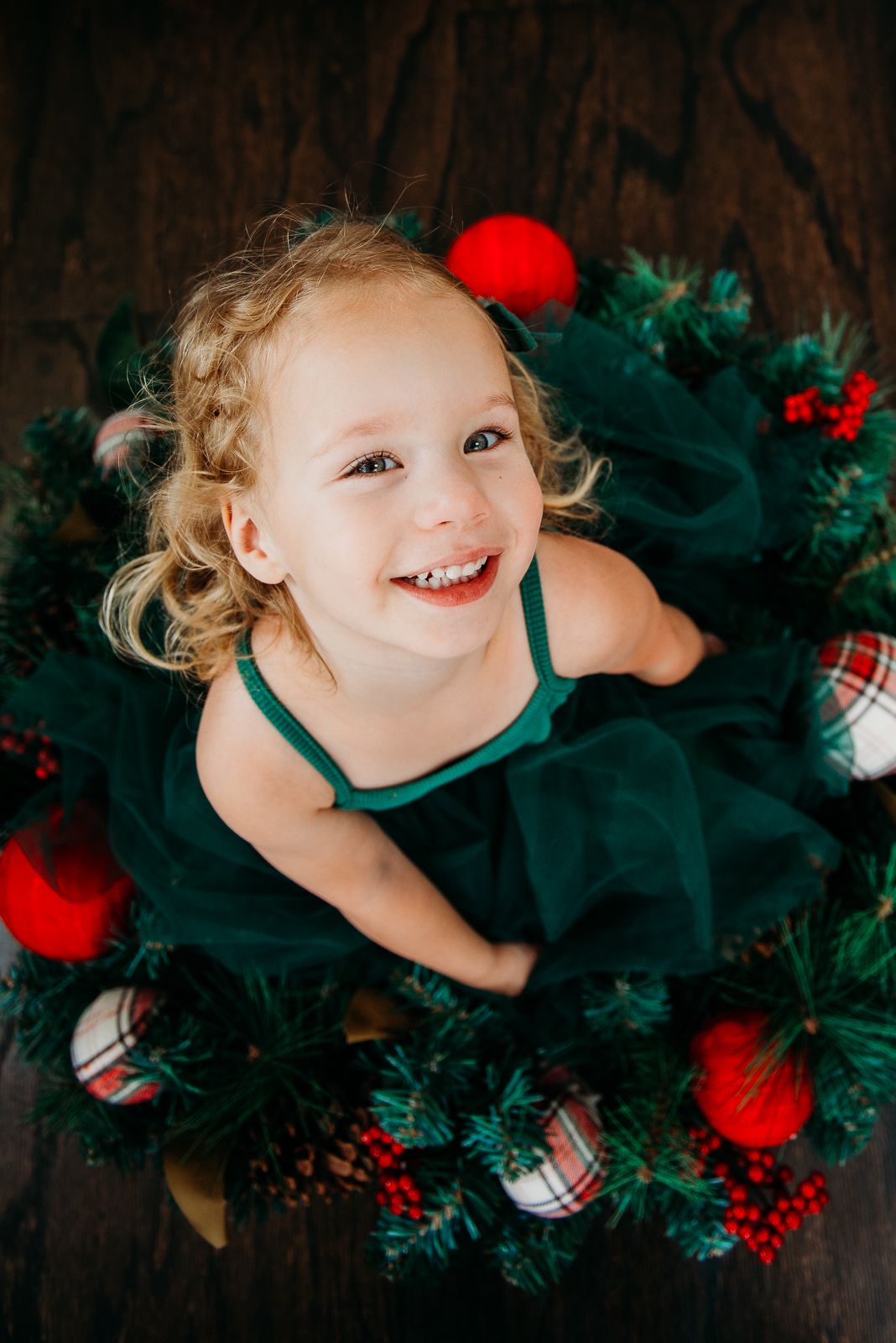 Young child in emerald green dress poses with Christmas wreath and decorations.