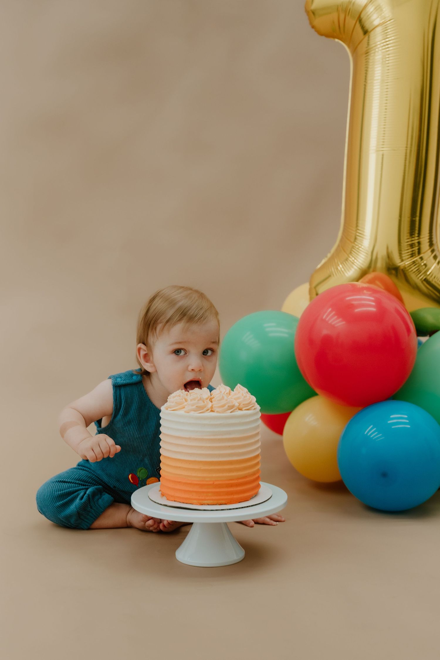 A first birthday cake smash photo series with colorful balloons and a white and orange ombre cake on a pedestal.