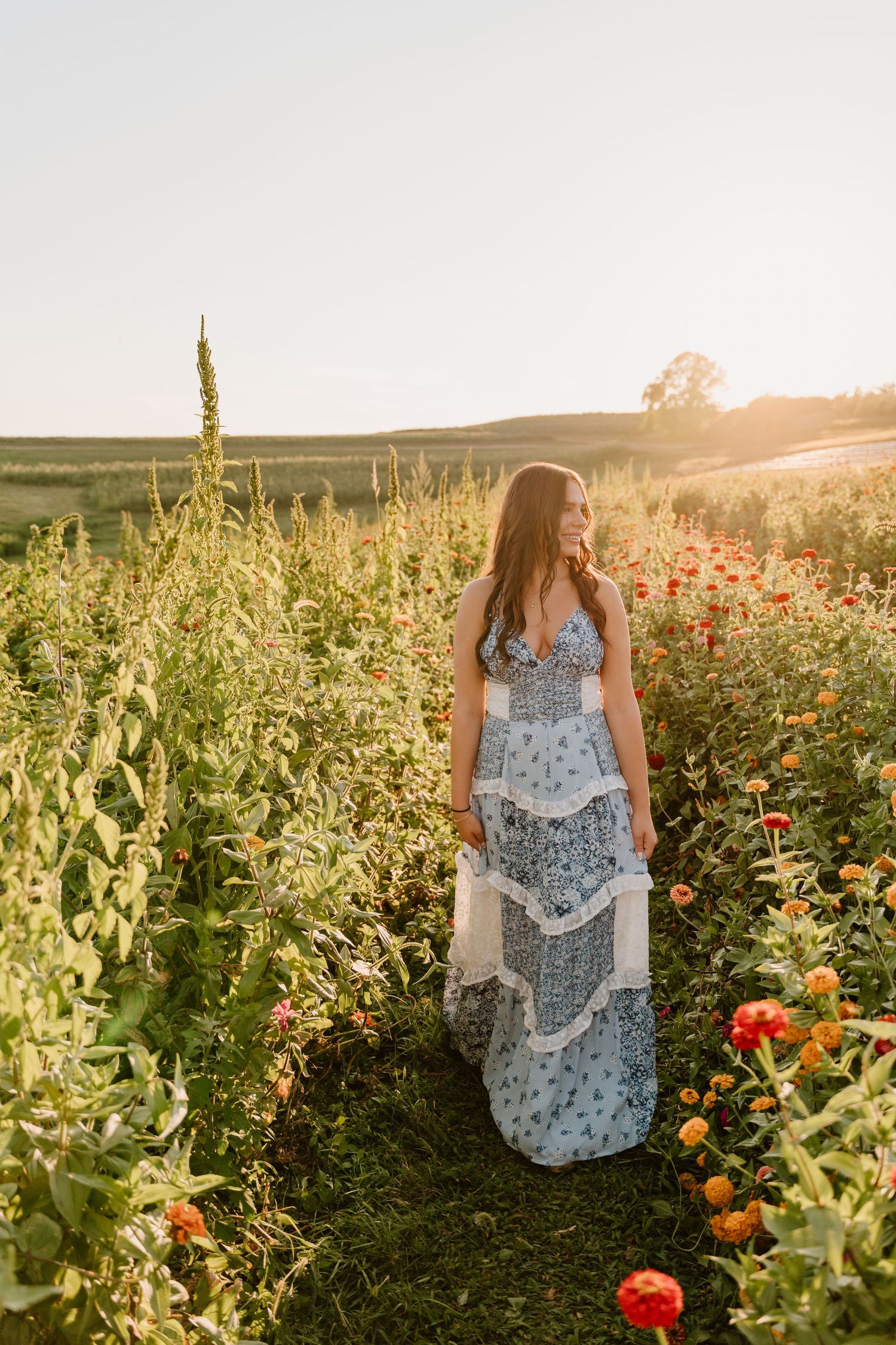 A person in a blue floral dress stands among vibrant wildflowers and tall plants in a golden sunset-lit field.