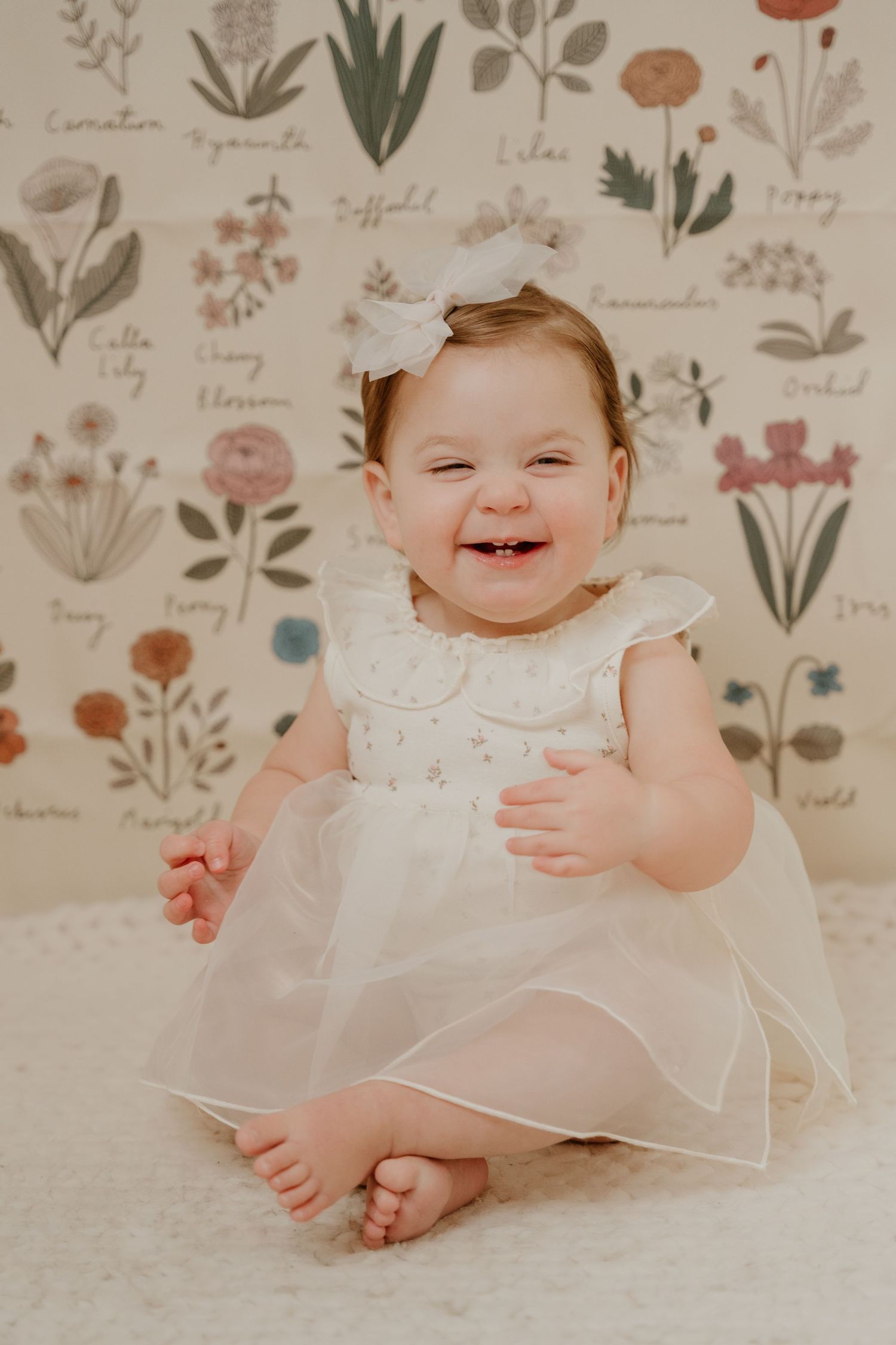 Baby wearing white dress smiles while sitting against floral patterned background.