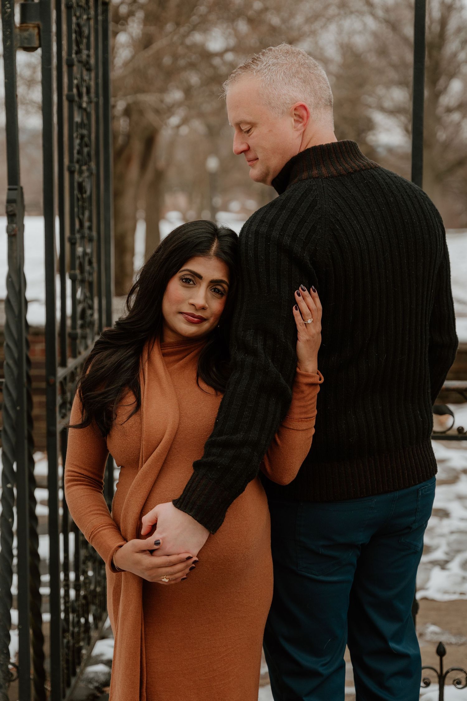 A couple poses together in winter, with the woman wearing a brown dress and the man in a black sweater by an iron gate.