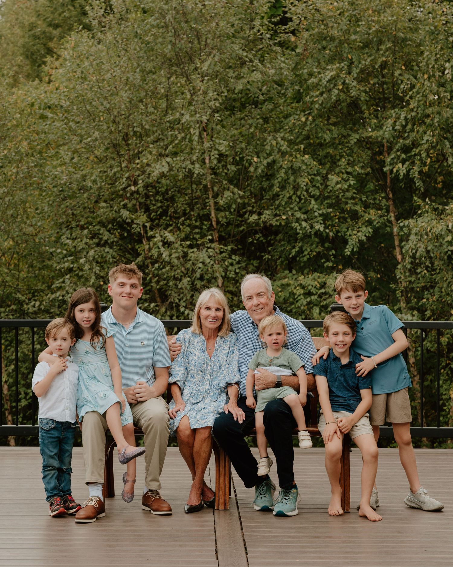 Multi-generational family sits together on wooden deck with lush green forest backdrop on sunny day.