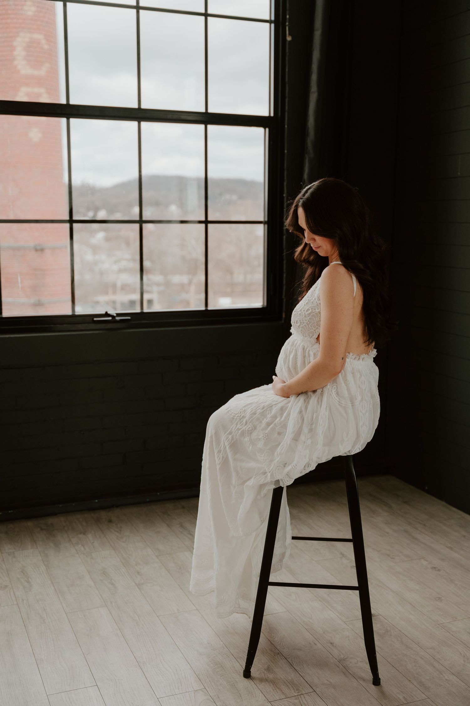Silhouette of a pregnant woman in a white dress sitting on a stool by a window during a moody maternity photoshoot.