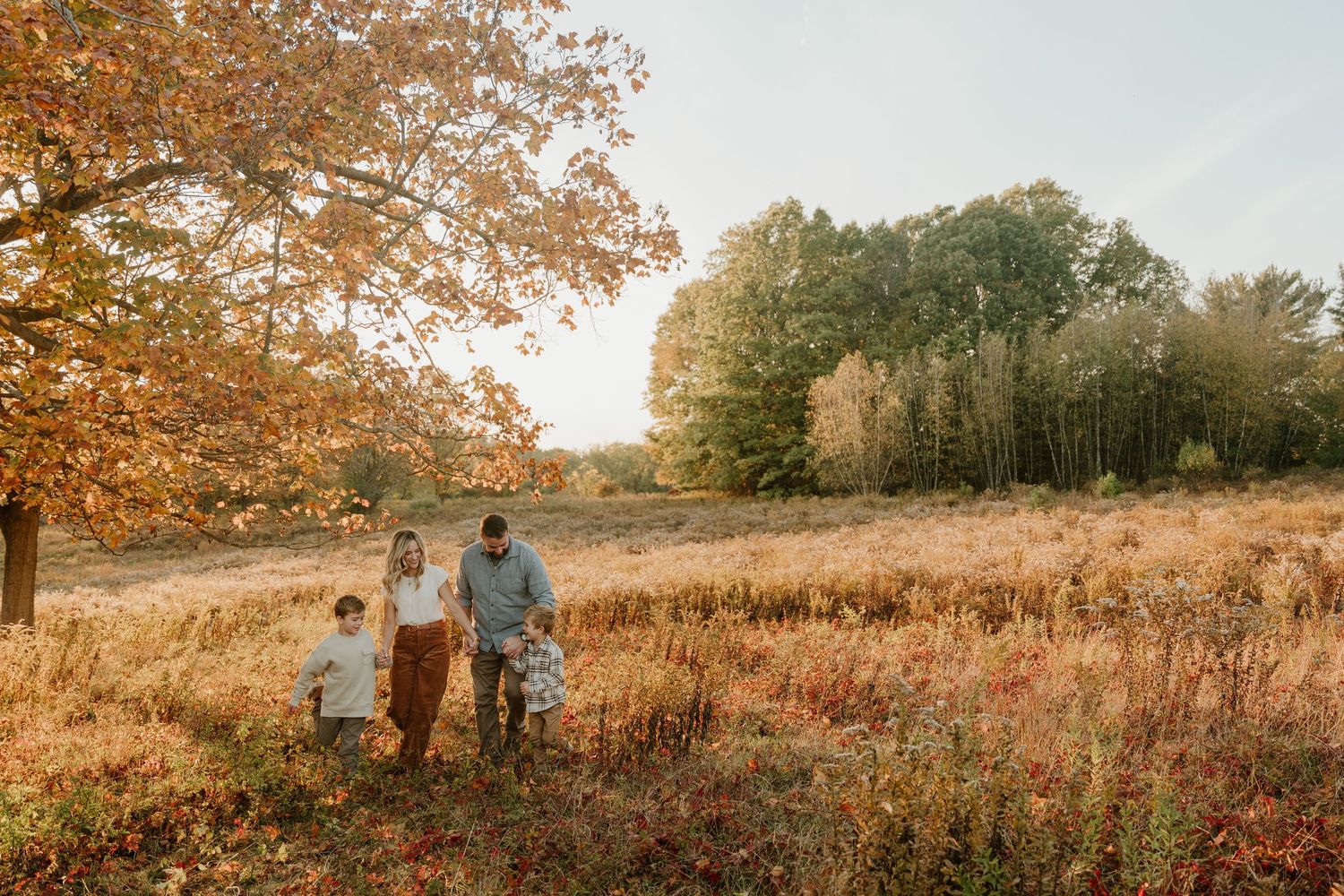 Couple walking through golden autumn field at sunset with tall grass and trees in warm tones.