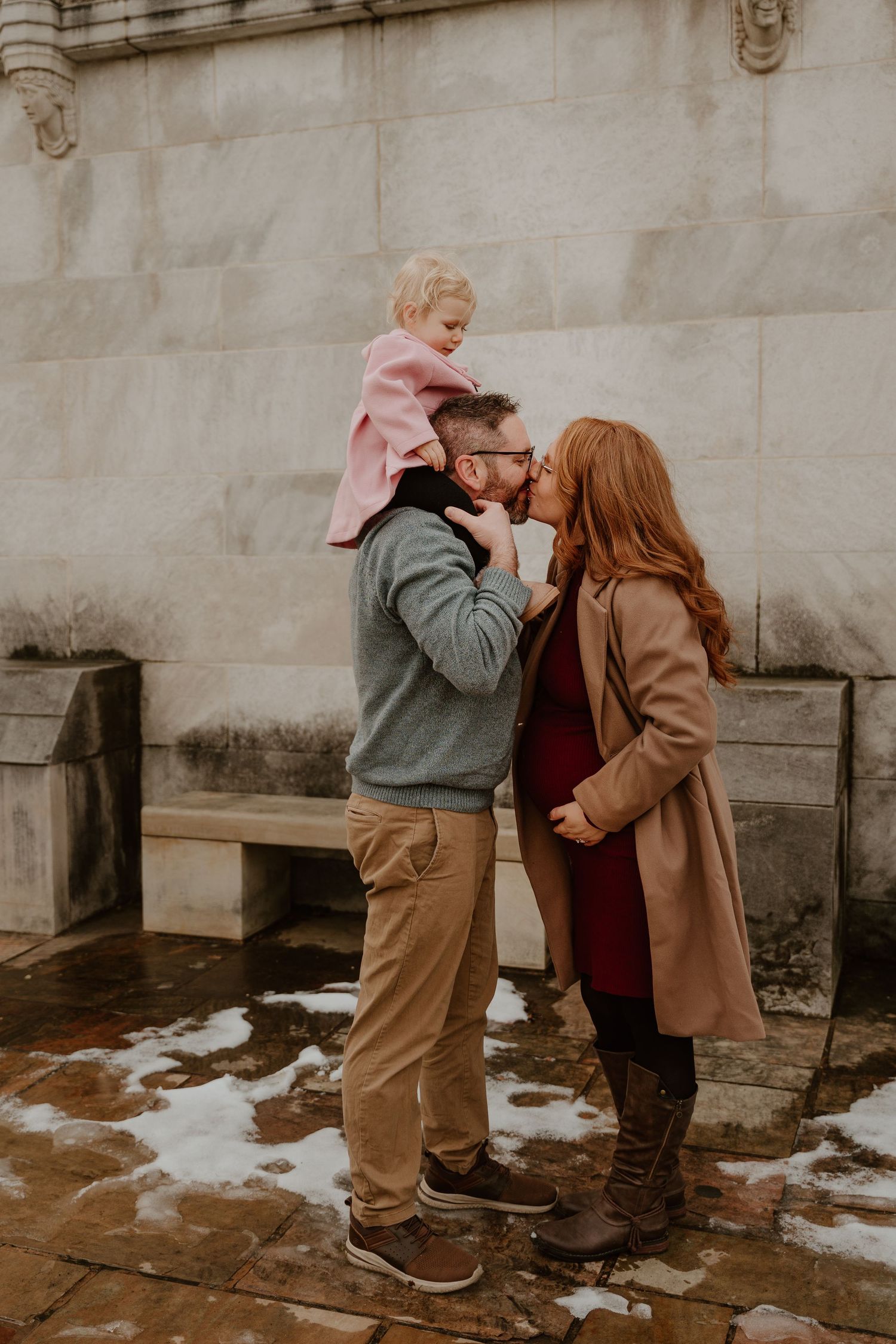 A family shares a tender winter moment together against a stone wall backdrop in warm coats.