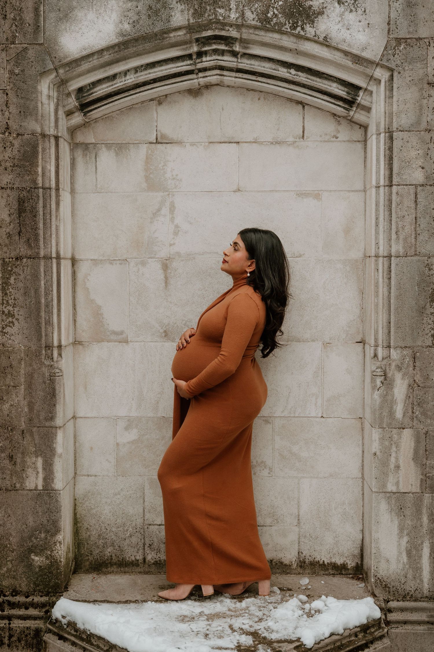 A pregnant woman in a brown maxi dress poses against a stone archway in a moody winter setting.